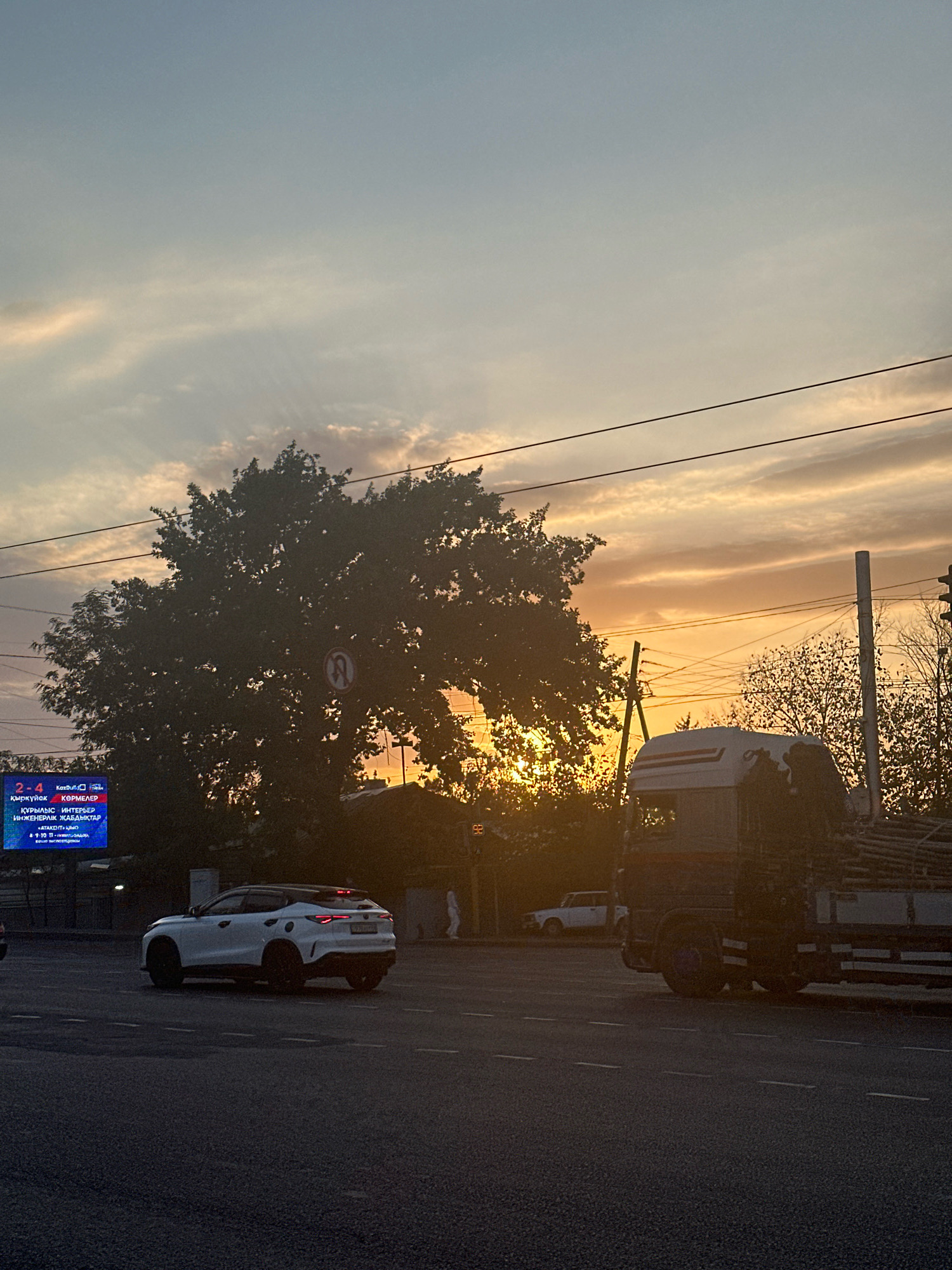The image depicts a street scene at dusk. A white SUV is visible on the road in the foreground, with a large truck loaded with lumber to its right. In the background, a large tree dominates the center of the frame, silhouetted against a sunset sky with orange and yellow hues. A digital billboard displaying text in a foreign language is also present to the left of the tree. Electrical wires crisscross the sky, and a traffic signal and various poles are scattered throughout the scene. The overall impression is one of an urban environment at the end of the day, with the natural beauty of the sunset contrasting with the man-made structures and vehicles.