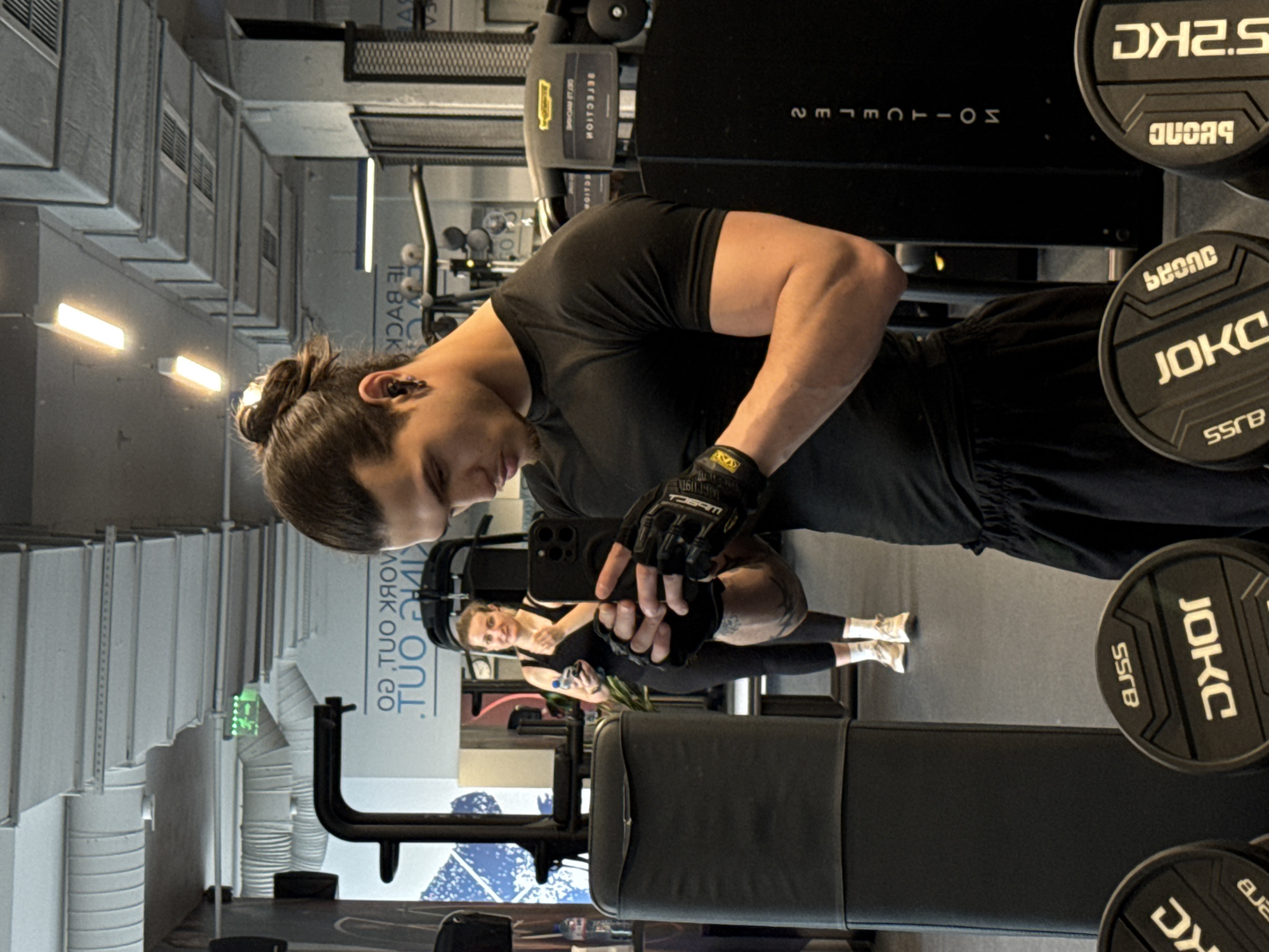 This indoor image captures a gym selfie, taken from a slightly low angle and originally rotated. The main subject is a man with dark hair tied in a bun, wearing a black t-shirt and fingerless weightlifting gloves. He holds a smartphone, appearing to take a mirror selfie, and his left arm displays a muscular bicep.
In the reflection behind him, a woman with light hair, dressed in a black athletic top and leggings with white sneakers, is visible. She appears to be smiling.
The setting is a modern gym or fitness center. Various gym equipment is present, including dumbbells with "JOKC", "55KB" (likely 5.5 KG), "DK2.5" (likely 2.5 KG), and "QUCAS" inscribed on them. A black exercise machine is visible in the background, showing reversed text like "TECHNOGYM" and "SELECTION" due to the reflection. The environment features industrial-style elements, such as exposed ventilation ducts, concrete walls, and bright rectangular light fixtures. A reflected sign in the background seems to partially read "WORK OUT Co" and "BE BACK". The overall mood is focused and active, typical of a workout session, under artificial lighting.