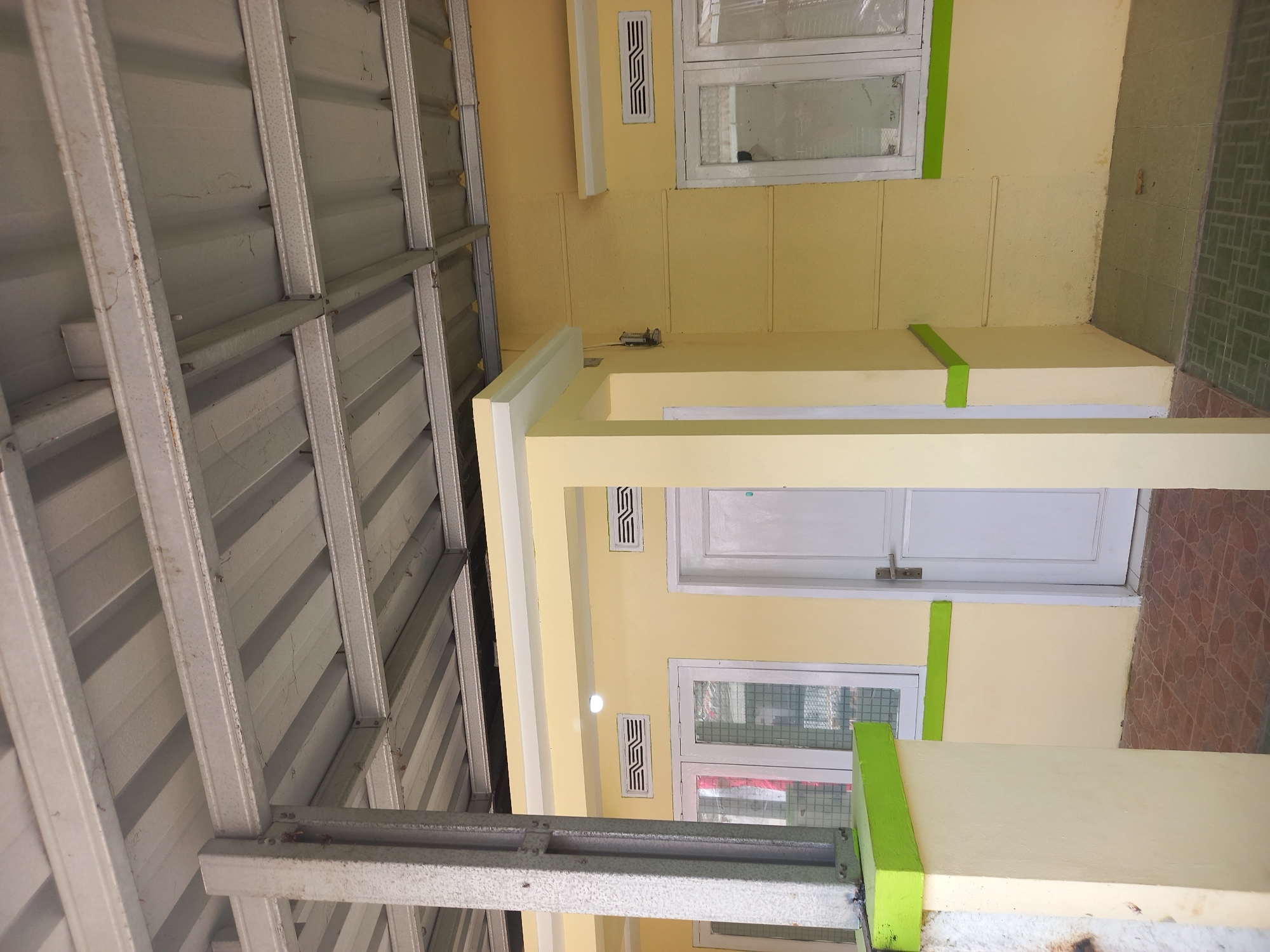 The image shows the exterior of a building with a yellow facade and a corrugated metal roof supported by a visible steel structure. The building features white-framed windows and a white door. There are also ventilation grilles on the wall. A concrete pillar with a green accent runs along the left side of the image, and the ground is paved with tiles. The scene is likely located in Cankuang, Indonesia, given the architectural style and the typical climate suggested by the open structure. No people are present in the image.