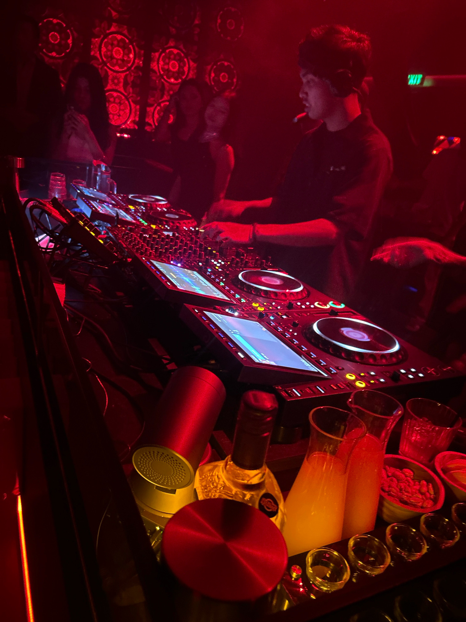 A DJ is working at a DJ booth in a nightclub. The DJ, a young male with headphones on, is standing and operating the equipment. He is smoking a cigarette. Several people are blurred in the background, mostly women. In the foreground, there is a bar with drinks, including bottles of alcohol and glasses filled with liquid, as well as small bowls containing snacks. The environment is dimly lit with red lighting and features decorative elements on the wall. The scene suggests a vibrant nightlife atmosphere.