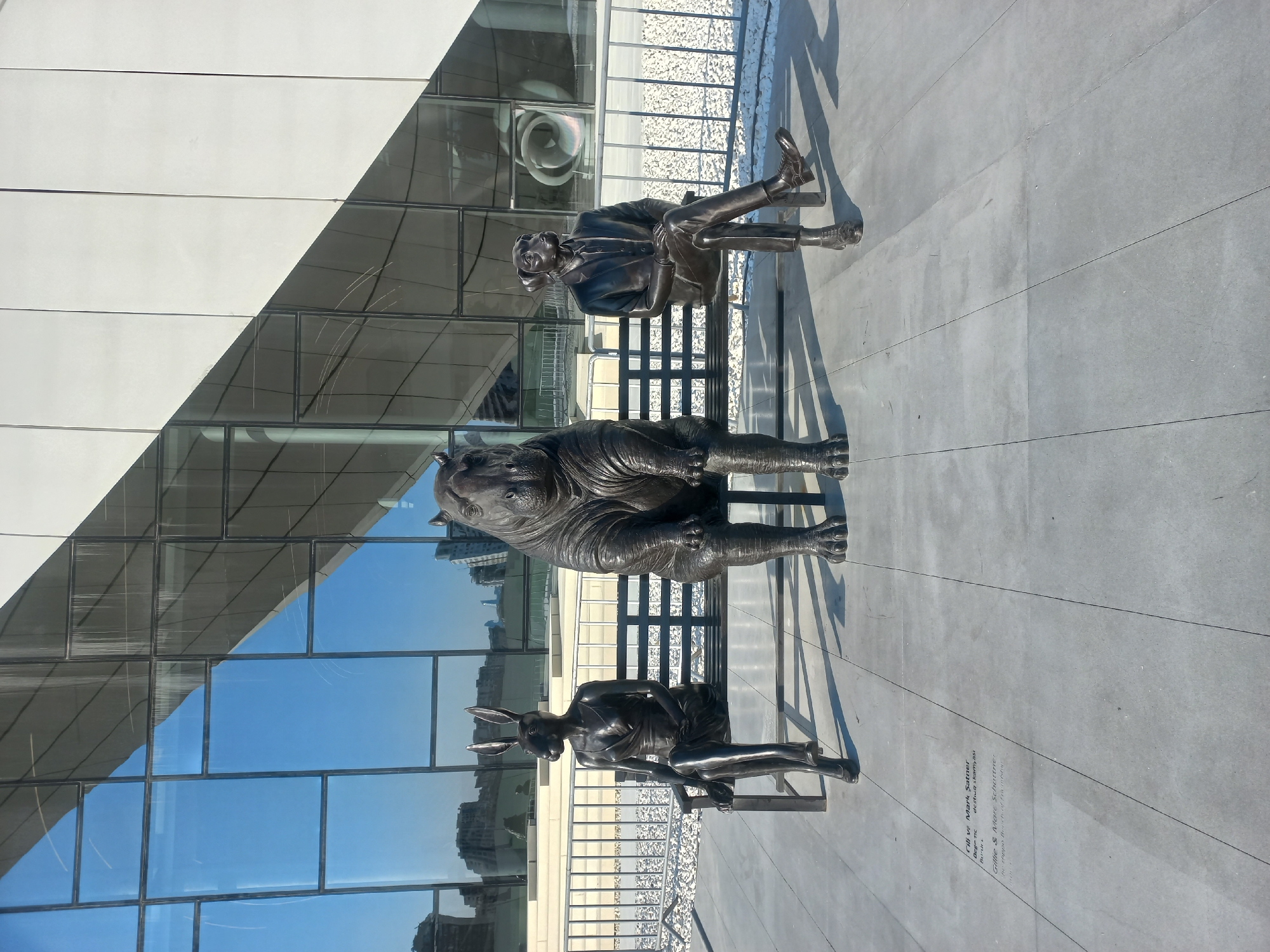 The image depicts a public sculpture installation located in Baku, Azerbaijan, likely outside the Heydar Aliyev Center, a building known for its distinctive fluid architecture. The sculpture features three bronze statues seated on a black bench. From left to right, the statues represent a rabbit-headed figure in a dress, a hippopotamus, and a dog-headed figure in a suit. The statues are life-sized or larger and are positioned close together on the bench. In the background, the sleek, modern facade of a building with large glass windows reflects the blue sky and surrounding cityscape. A metal fence and white gravel are visible in the mid-ground. The ground in front of the bench is paved with large square tiles. There are inscriptions on the ground in front of the bench, though they are difficult to read clearly. The lighting suggests it is a bright, sunny day.