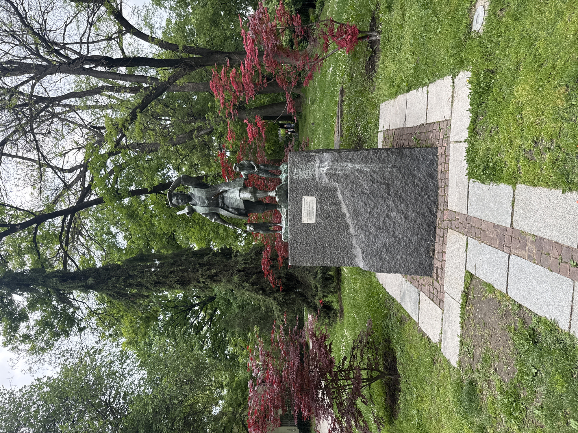 The image depicts a statue of a historical figure, likely of importance to Plovdiv, Bulgaria. The statue itself features a man in period attire, possibly armor, and appears to be made of bronze or a similar metal. It is mounted on a dark gray, stone pedestal with a plaque attached to its side. The pedestal is surrounded by a pathway of light gray stone pavers, bordered by smaller, possibly brick, elements. The pathway is laid upon a lawn of green grass.
The backdrop of the statue consists of lush greenery, including tall trees with expansive canopies. A tree with reddish-purple leaves is also visible near the statue, adding a touch of color to the scene. The environment suggests a park or public garden.
Given the subject matter, the statue likely stands within a historic or culturally significant area of Plovdiv.