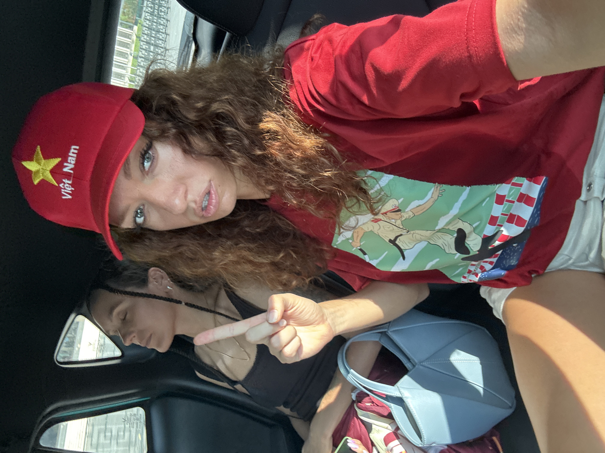 A sideways, medium-shot photograph shows two women inside a car. The woman in the foreground is light-skinned, with curly brown hair and wearing a red baseball cap with the words “Việt Nam” and a yellow star embroidered on it. She wears a red t-shirt over a printed t-shirt with an illustration of a person and an American flag. The woman is pointing toward the other woman.
The other woman, who is dark-skinned, is in the background. Her eyes are closed and her face is oriented toward the left of the frame. She is wearing a black tank top. A window is behind her.
A light blue handbag sits in the foreground. The location of the photograph is unspecified. Hà Nội, Vietnam is not recognizable in the image.