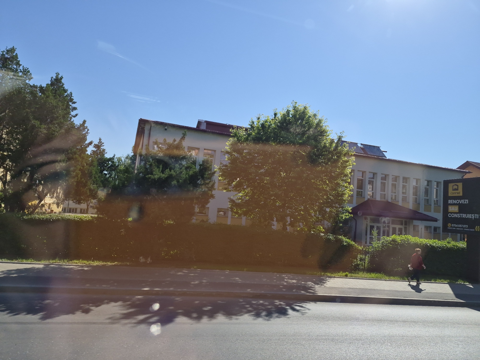 The image shows a street scene in Fălticeni, Romania. A building with a red roof and many windows is visible, along with trees and a sidewalk. A person in a red jacket is walking on the sidewalk. A billboard with Romanian text and a phone number is also present. The sky is clear and blue, and the sun is shining. The image appears to have been taken from a vehicle, as evidenced by reflections on the glass.