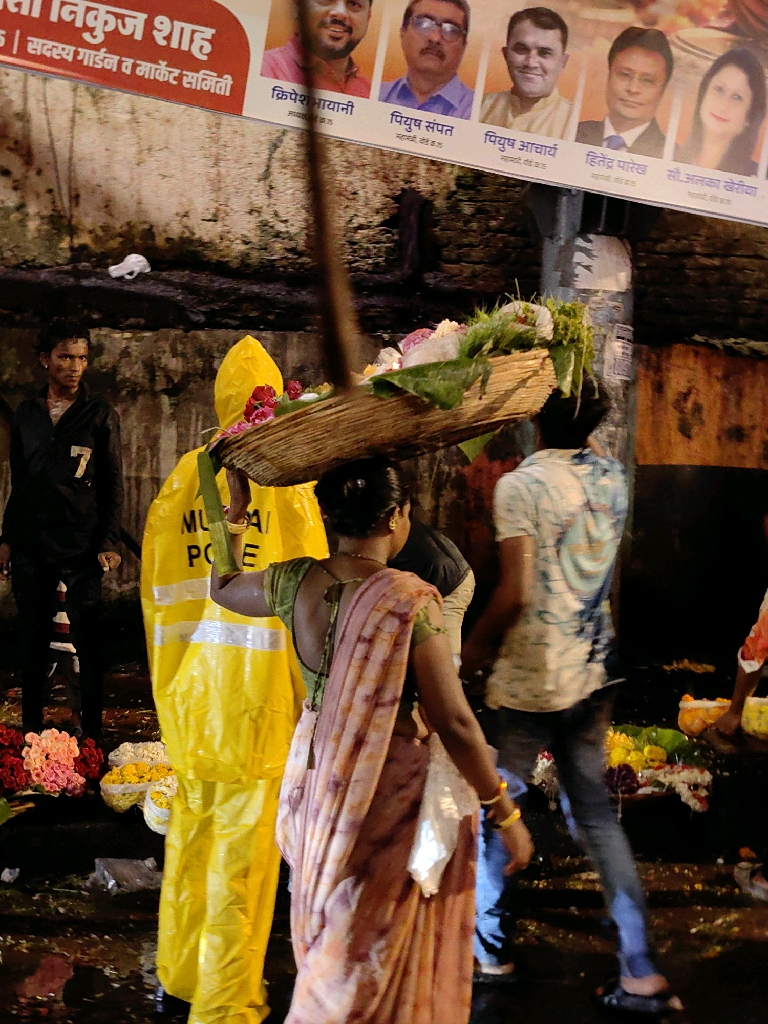 The image depicts a street scene in Mumbai, India, likely at night or during twilight. The foreground features several individuals and elements related to a street market, specifically focusing on flowers.
On the left, a young man wearing a black shirt with the number "7" printed on it stands looking to the side. He is dressed in black pants.
To his right, a person in a bright yellow, full-body rain suit with reflective silver stripes is visible from the waist down. The suit has "MUMBAI POLICE" printed on it in black capital letters.
In the center of the frame, a woman, from the back, is carrying a woven basket filled with flowers and greenery on her head. She is wearing a pink and orange sari and a green blouse. Her left arm is bent, and she is wearing a yellow bangle.
Behind the woman and to the right, a man in a white t-shirt with a blue and white abstract print is walking away from the viewer. He is wearing dark blue jeans.
In the background, a large banner with portraits of several men and women is displayed. The text on the banner is in Marathi and appears to be related to a local committee or organization. Below the banner, a brick wall forms the backdrop.
The ground is wet and reflective, suggesting it has recently rained or is damp. Several baskets of flowers, including red, pink, yellow, and white varieties, are visible on the ground, indicating a flower stall. The overall impression is a busy, perhaps slightly damp, market atmosphere.
