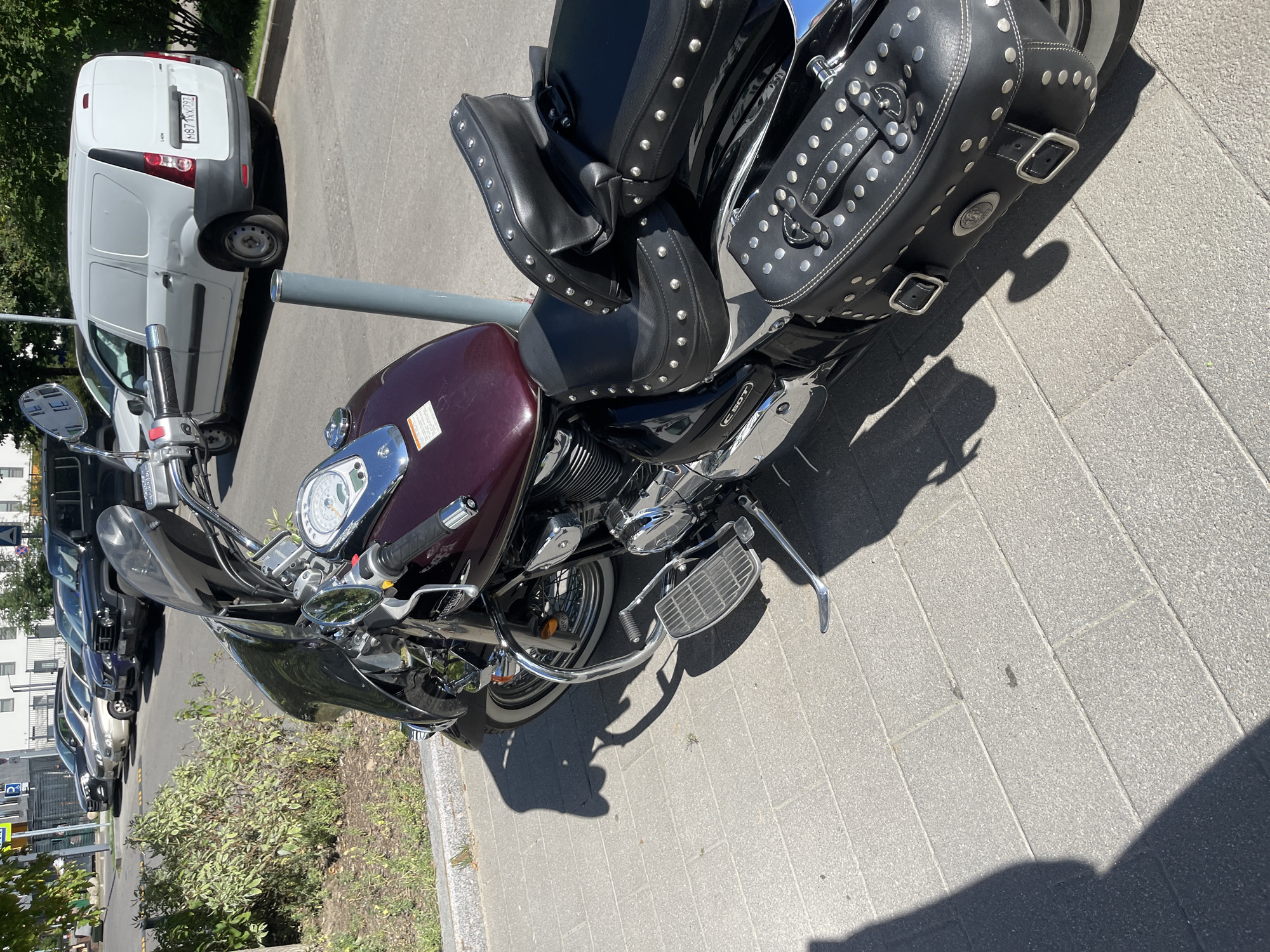 In Kommunarka, Russia, on a paved sidewalk next to a street with parked cars, a dark red and black motorcycle is parked. The motorcycle is a cruiser style with a large windshield, chrome accents, and studded black leather saddlebags and seats. The motorcycle's handlebars are visible, as is part of the engine and exhaust system. Sunlight casts shadows of the motorcycle and a person onto the sidewalk. In the background, several cars and a white cargo van are parked on the street. Buildings and trees are visible further in the background.