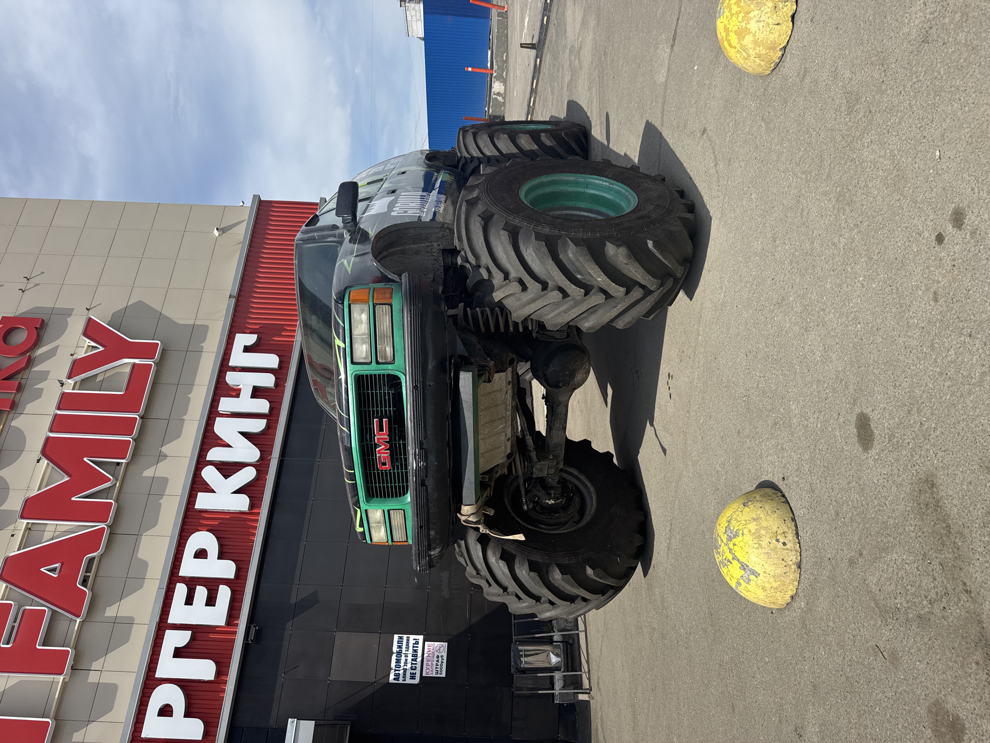 The image depicts a modified, raised GMC truck parked in front of a "Burger King Family" restaurant in Izhevsk, Russia. The restaurant's sign is in red and white, and there is a sign in Russian prohibiting parking within 20 meters of the building and also prohibiting smoking. The truck has oversized tires with green rims. Two yellow parking bollards are visible in front of the truck. The sky is blue with some clouds.