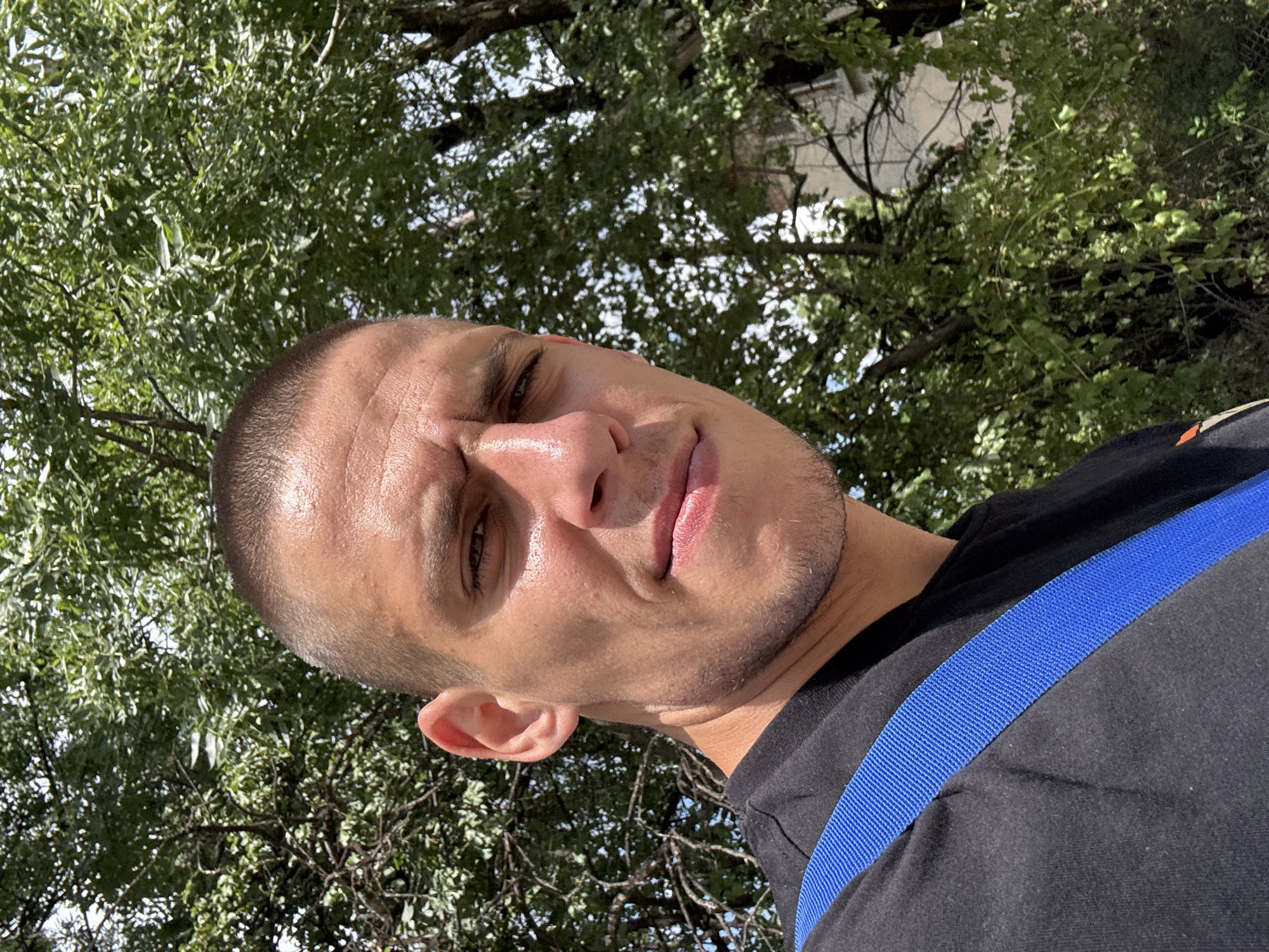 A young man with a buzz cut, wearing a black t-shirt and a blue strap across his chest, looks directly at the camera with a slight squint. He appears to be outdoors, with lush green trees and foliage forming the backdrop. A building with windows is visible in the background, partially obscured by the trees. The lighting suggests it is daytime, possibly a sunny day. The image composition is a close-up, focusing on the man's face and upper torso. The man's skin appears to be sweating. The location is identified as Sofia, Bulgaria.