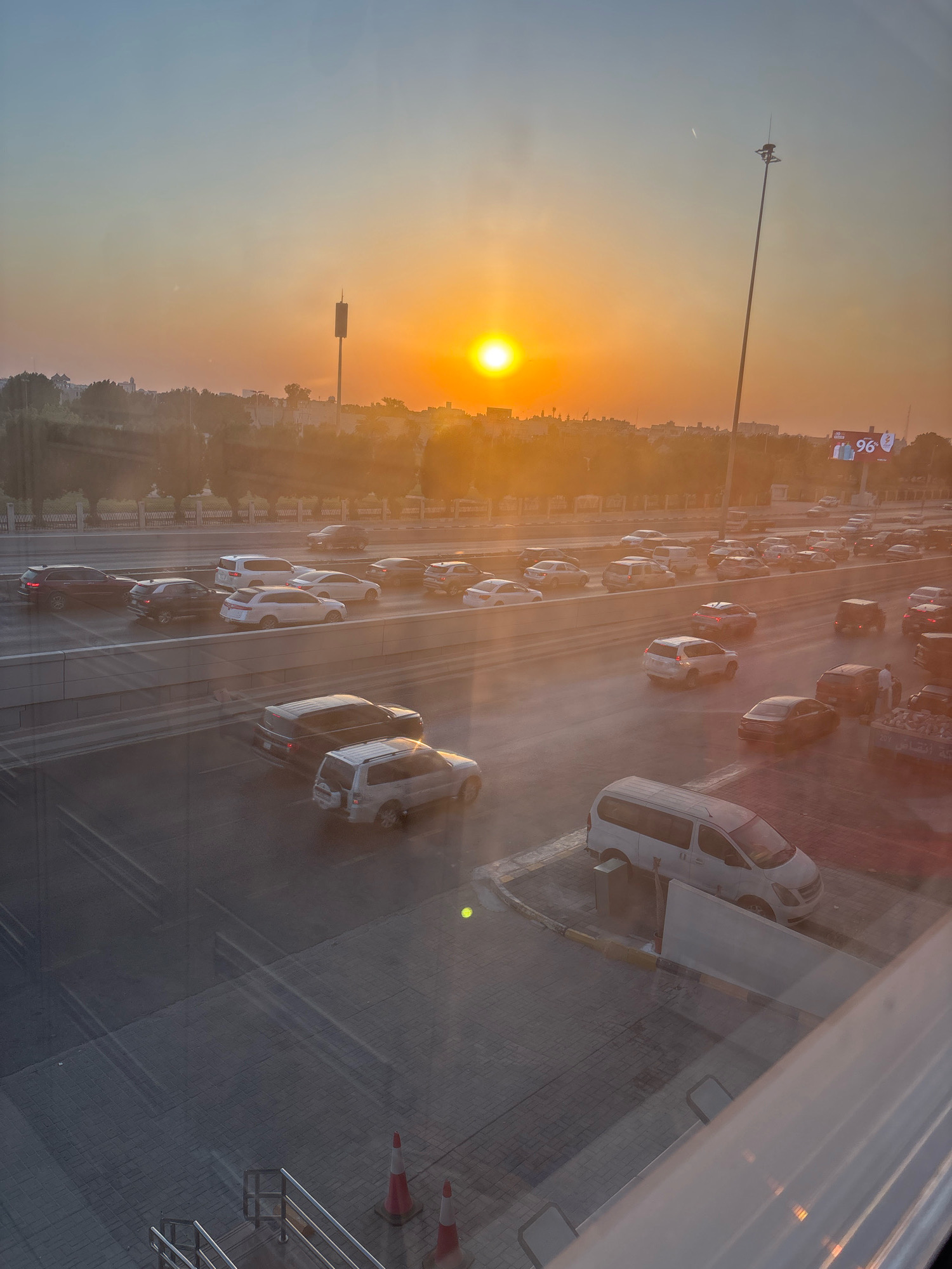 This is a photograph taken at sunset in Dhahran, Saudi Arabia, showing a busy highway with a significant amount of traffic. The sun is low on the horizon, casting a warm, orange glow over the scene and creating a hazy atmosphere. The highway is multi-laned, with cars, SUVs, and vans driving in both directions. Some of the vehicles appear to be stationary or moving slowly, suggesting a traffic jam. In the background, there are buildings and trees, and a tall communication tower is visible on the left. A large digital billboard displaying the number "96" is positioned above the highway on the right side. The foreground shows a section of a street or sidewalk with traffic cones and a handrail, suggesting the vantage point is from an elevated position, possibly a building overlooking the highway. The lighting and reflections on the glass indicate the photo was taken from indoors.