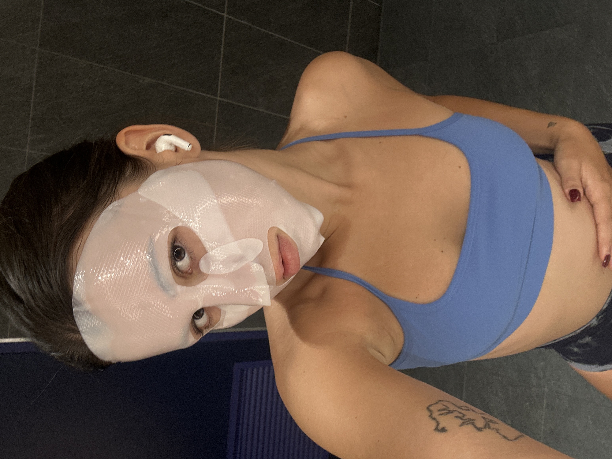 A woman with a sheet mask on her face, wearing a blue sports bra and dark patterned shorts, is shown from the chest up in a medium shot, looking at the camera. She has dark hair pulled back and is wearing white wireless earbuds. A tattoo is visible on her left arm. The background is dark with a tiled wall and what appears to be a blue slatted door or wall to the left. The lighting is dim, suggesting an indoor setting. There is no discernible location in Si Sunthon, Thailand.