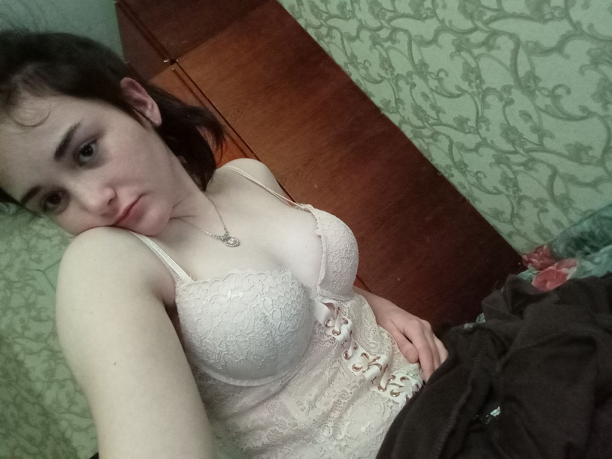 A young woman with dark hair is lying down, wearing a white lace bustier and a silver necklace. She is looking at the camera with a neutral expression. The background includes a light green patterned wall and a dark wood cabinet. Part of a dark-colored garment is visible at the bottom right. No details indicate this image was taken in Poltava, Ukraine.