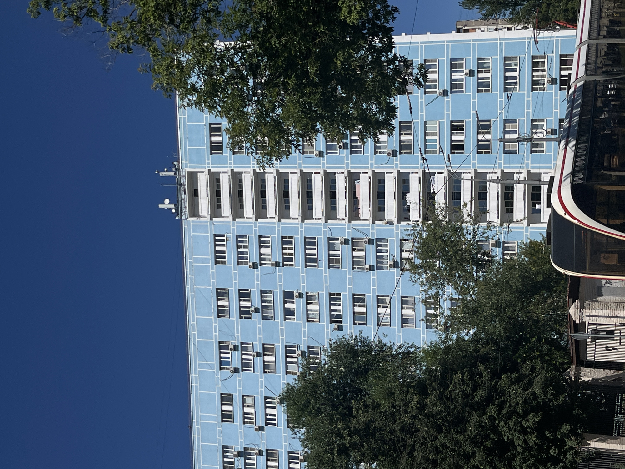 This media file depicts a tall, light blue building with a repeating pattern of windows and horizontal white lines on its facade. The building appears to be a residential or office building, typical of urban architecture. Many windows have white shutters, some open and some closed, with some also featuring visible air conditioning units mounted below them. The building is partially obscured by the lush green foliage of trees in the foreground, suggesting a warm, sunny day with clear skies, as indicated by the deep blue of the sky.
On the right side of the frame, a portion of a modern bus or tram is visible, with its distinctive red and white striped livery and dark tinted windows. The text "WWW.WWW" is visible on the side of the vehicle, likely part of an advertisement or branding. Several aerials or antennas are mounted on the rooftop of the building, hinting at its function or the presence of communication equipment. Power lines and cables are also visible, crisscrossing in front of the building, a common sight in urban environments.
The perspective suggests the photo was taken from a low angle, looking up at the building. The overall scene conveys a sense of everyday urban life in Moscow, Russia, under a bright sky, with the presence of both modern infrastructure and natural elements like trees. No people are clearly visible, and the focus is on the architectural elements and the surrounding environment.
