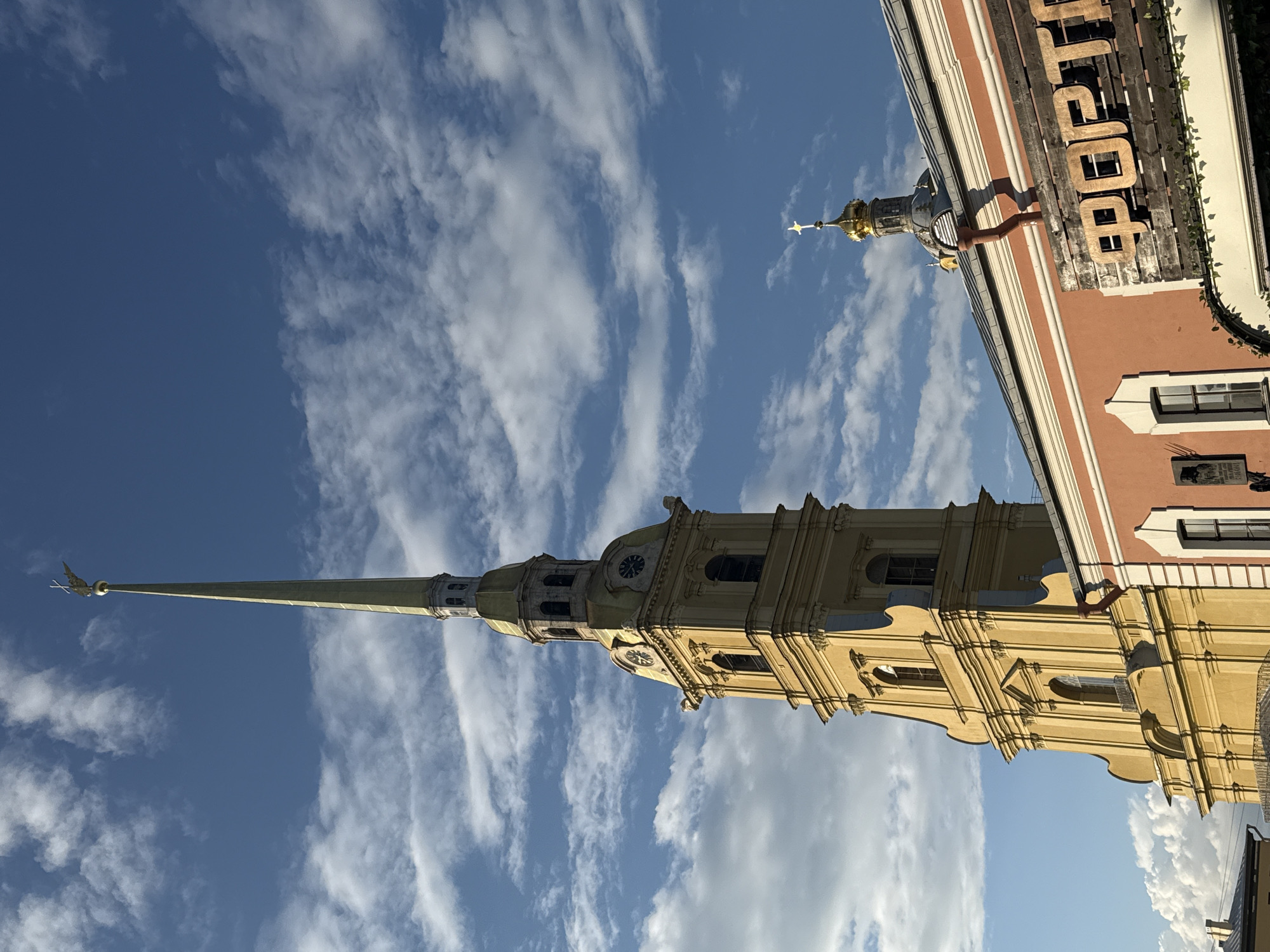 This media file captures a low-angle shot of a tall, slender spire, likely belonging to a church or cathedral, against a bright blue sky dotted with wispy white clouds. The spire is predominantly green with a golden finial at its apex. Below the spire, the upper section of a yellow building is visible, featuring architectural details such as windows, decorative moldings, and what appear to be clock faces. To the right, a portion of an adjacent building with a salmon-pink facade is also in view, showcasing its windows and some decorative trim. The image is oriented vertically, suggesting it was taken looking upwards. No people are visible, and no specific activity or event is depicted. The lighting suggests it is daytime, possibly late afternoon given the warm tones on the yellow building. The presence of distinctive architecture, particularly the spire, strongly hints at a European, and specifically, a Russian or Saint Petersburg setting, as suggested by the provided location context. There is some visible text on the salmon-pink building, which appears to be a wooden sign with large, dark letters, possibly spelling out a name or word, but it is partially obscured and difficult to read clearly. The overall mood is one of architectural grandeur and a pleasant, clear day.