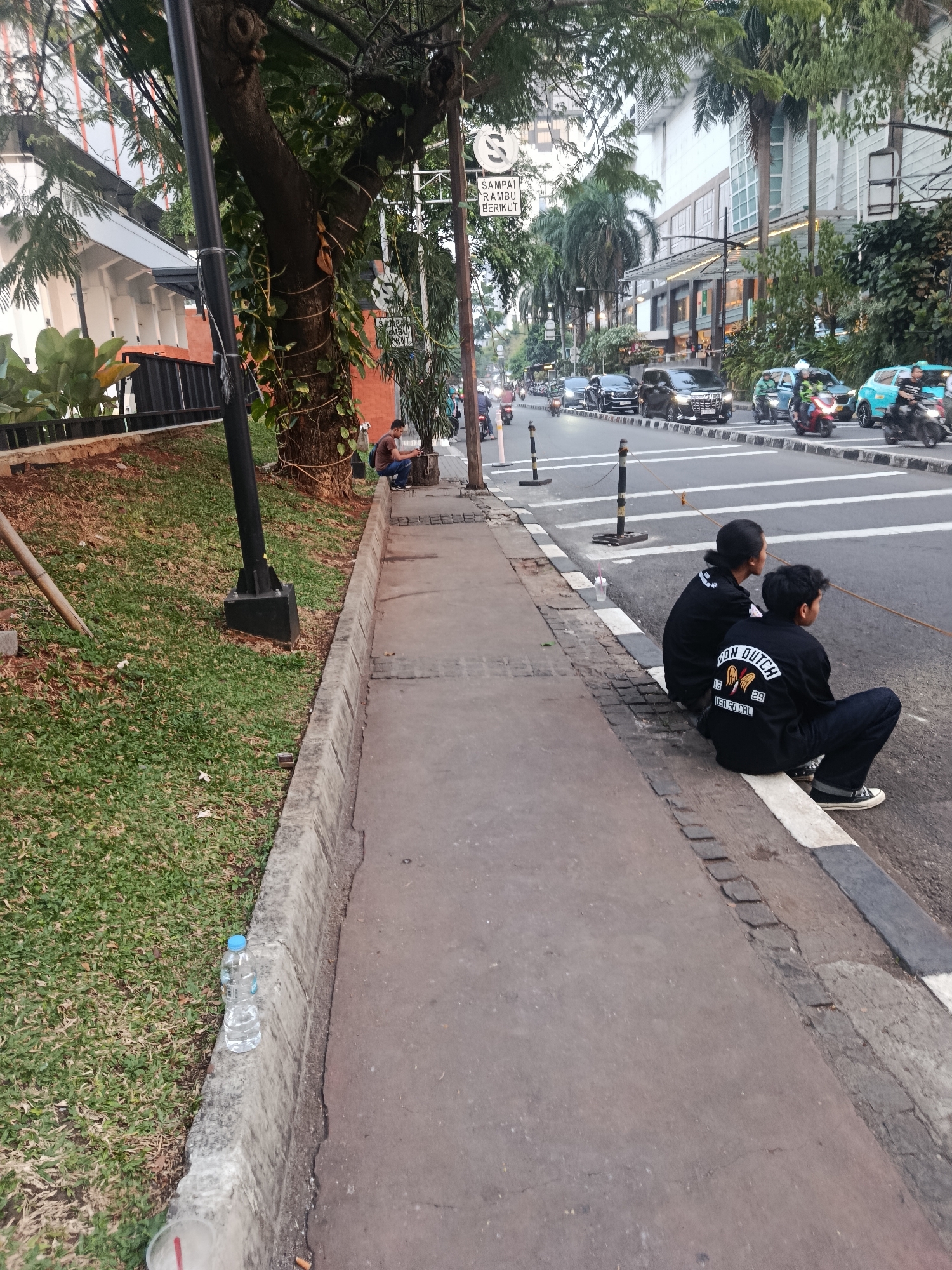 The image depicts a street scene in South Jakarta, Indonesia, with a focus on a sidewalk and the adjacent road. Two individuals, likely young men, are sitting on the curb on the right side of the frame, facing the road. They are both wearing black jackets with white lettering on the back. The jacket of the person closer to the camera appears to have "NON DUTCH" and "USA. SO CAL" printed on it.
The sidewalk is paved with concrete and has a curb separating it from a grassy area on the left. A lamppost stands on the grassy area. Further to the left, a tree with vines wrapped around its trunk is visible. Beyond the sidewalk and grass, a building with modern architecture and glass windows is seen, lined with palm trees.
The road in front of the curb is busy with traffic, including cars, motorcycles, and what appears to be a ride-hailing vehicle. The road is marked with white lines and a pedestrian crossing. A traffic sign with a "S" symbol and Indonesian text is visible in the background. A plastic bottle of water sits on the curb in the foreground on the left. The scene appears to be in an urban environment, possibly a commercial or residential area, during daylight hours.
