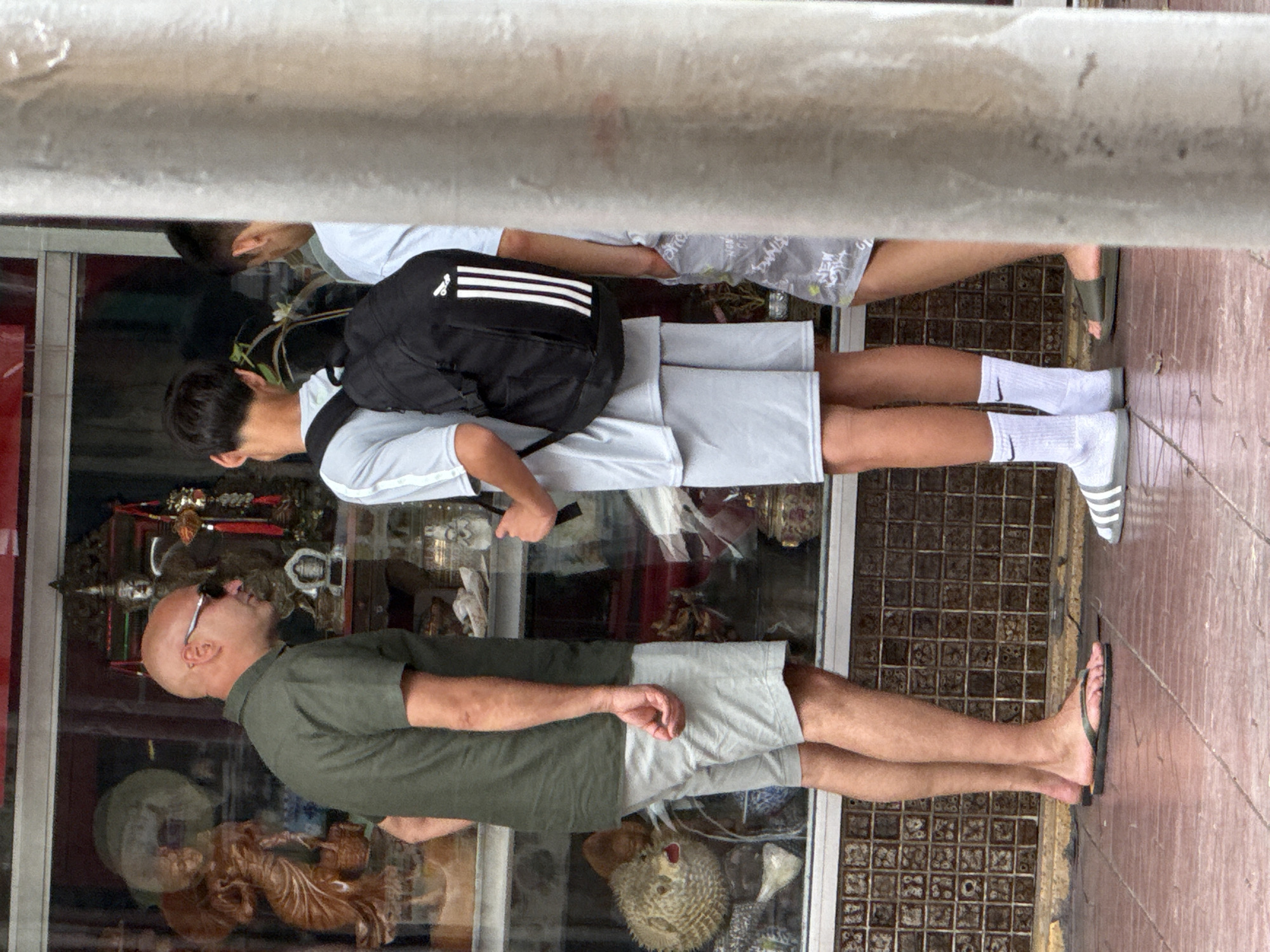 This is a full shot of three individuals standing on a tiled sidewalk in front of a shop window displaying various items. The shop window has a tiled border.
The person on the left is a bald man wearing sunglasses, a green short-sleeved collared shirt, and light gray shorts. He is standing with his left hand behind his back and his right hand in his pocket.
The person in the middle is a younger individual, likely male, wearing a light gray t-shirt with white stripes on the sleeves, and light gray shorts. They have a black backpack with white stripes on it, worn over their shoulders.
The person on the right is partially obscured by a concrete pillar. They appear to be wearing a light-colored shirt and gray shorts with a graphic print. They are standing with their right hand in their pocket.
The shop window contains various items including figurines, a pufferfish, and decorative jars. The sidewalk is made of reddish-brown tiles arranged in a grid pattern. The background appears to be a street scene. The overall lighting suggests it is daytime.