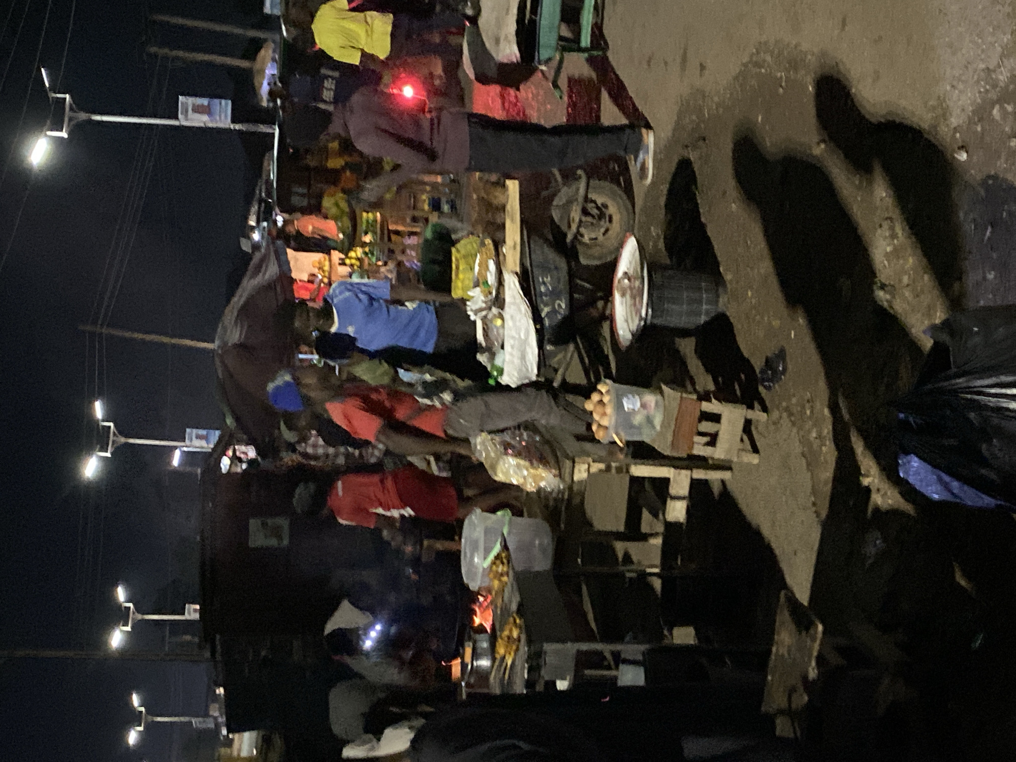 The scene depicts a bustling street in Bukuru, Nigeria, at night. The area is illuminated by solar-powered streetlights. Several people are visible, engaged in various activities. A makeshift market is set up along the street, with vendors selling goods and cooked food. Evidence of a small fire indicates food preparation. The ground is rough, with visible shadows and debris. A cart or vehicle with a spare tire is partially visible, suggesting mobile commerce.