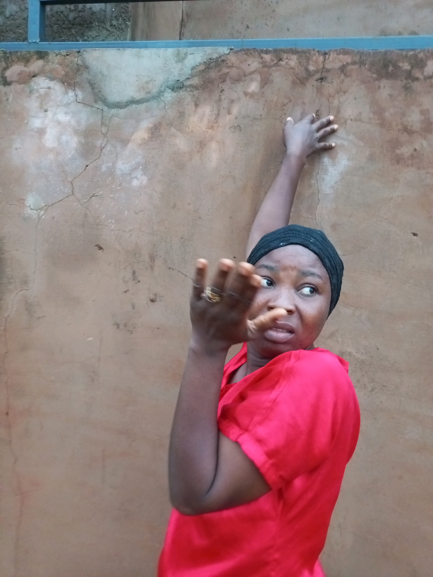 A medium shot shows a Black woman wearing a red top and a black headscarf. She is standing in front of a cracked, tan-colored wall and appears to be posing with her left arm raised and bent at the elbow, hand extended forward, with fingers splayed. Her right arm is bent, with her right hand resting on her right shoulder. She is looking to her right, with a surprised or concerned expression on her face. A blue railing is visible along the top of the frame. The image appears to have been taken outdoors, possibly in a courtyard or alleyway. The background wall is textured and shows signs of age and wear. The lighting suggests it is daytime, with shadows cast by the woman. There are no other people or objects visible in the frame.