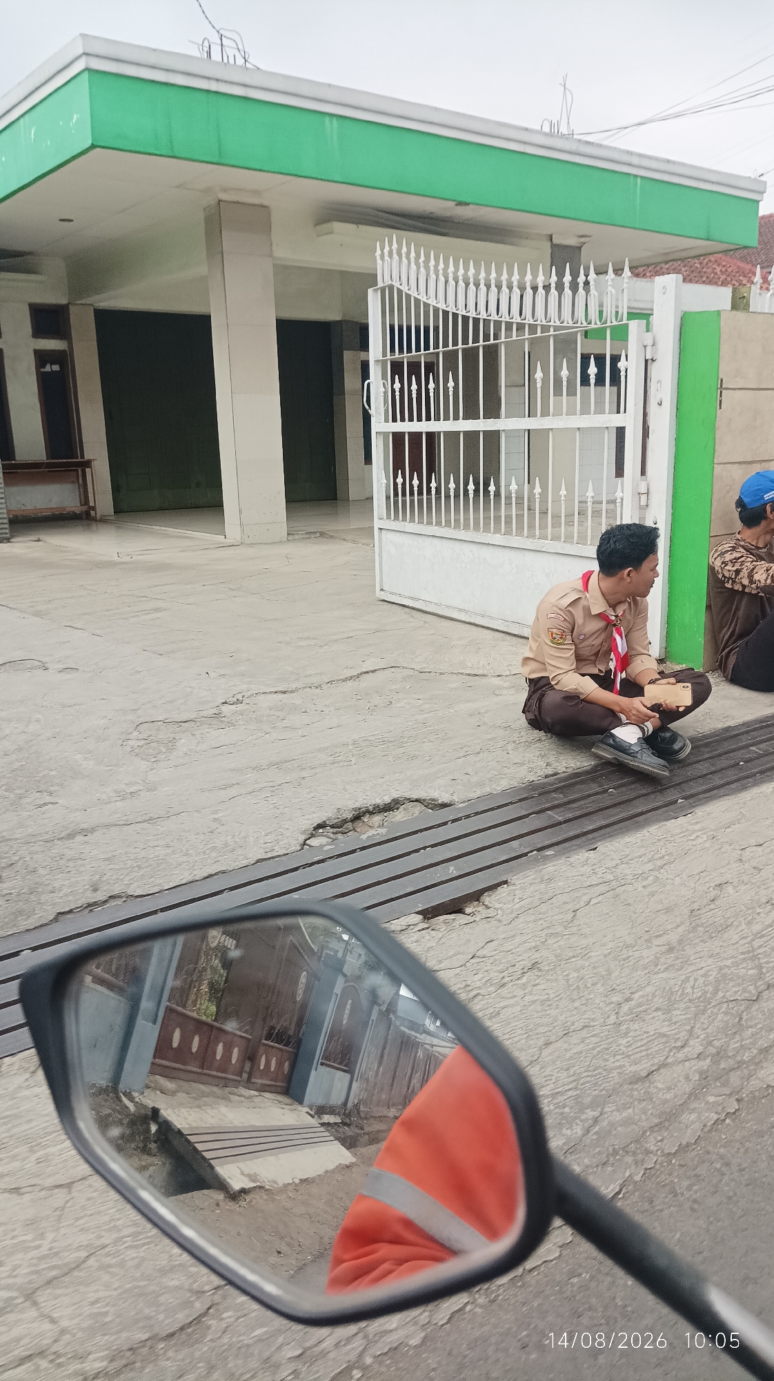 The image captures a scene on a concrete pavement in front of a building with a green overhang. A young man in a scout uniform, consisting of a tan shirt with badges and brown pants, is sitting cross-legged on the ground, holding a small brown object, possibly a book or phone. He is looking to his right. To his right, another person is partially visible, wearing a brown patterned shirt and a blue cap. The foreground of the image is dominated by a motorcycle's side mirror, reflecting a house with a blue facade and a brown gate, and a person wearing an orange jacket. The timestamp "14/08/2026 10:05" is overlaid on the bottom right corner of the image. The environment suggests an urban or semi-urban setting in Tasikmalaya, Indonesia, with the architectural style of the building and the presence of a motorcycle being common in the region. The pavement shows cracks and some wear, indicating it's a public outdoor space.