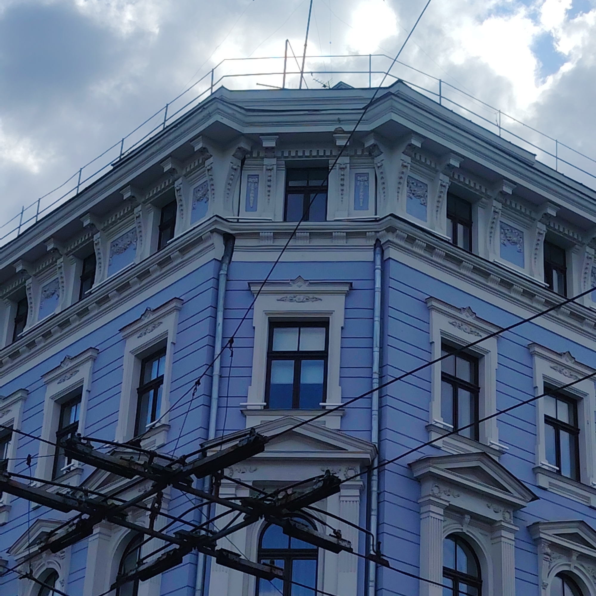 The image shows a close-up, low-angle view of the corner of a blue, ornate building with white trim. The building has multiple windows with dark frames, some rectangular and some arched. There is decorative architectural detailing around the windows and along the roofline. Overhead, a complex network of dark electrical wires and supporting structures crisscrosses the frame, obscuring parts of the sky and the upper portion of the building. The sky is partly cloudy. The building appears to be located in the city of Riga, Latvia, based on the context provided. There are no people or animals visible in the image. The overall impression is one of an urban architectural scene with a strong emphasis on the building's facade and the overhead infrastructure.