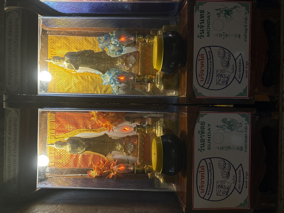 The scene depicts an interior view within a religious establishment in Ban Hin Sam Kon, Thailand. Two illuminated, glass-fronted wooden display cases stand side-by-side, each dedicated to a specific day of the week for offerings.
The left display case, associated with "SUNDAY," contains a standing, gold-colored Buddha statue with a pointed finial. The statue's right hand is raised in a gesture of blessing, and it is positioned against a textured, reddish-orange patterned fabric background. Artificial orange flowers flank the statue. Two flame-shaped electric candles emit an orange glow from the base, supplemented by a white light bulb above. Below this case, a black offering bowl with a yellow lid rests on a wooden shelf. A white sign with green borders identifies the day as "วันอาทิตย์" (Sunday) and "SUNDAY," displaying illustrations of banknotes and coins, a phone number (081-0504464), and a depiction of the Buddha posture "ปางถวายเนตร กำลัง 6" (Pang Thawai Net, strength 6).
The right display case, associated with "MONDAY," is similar in structure. It houses another standing, gold-colored Buddha statue, also with a pointed finial and its right hand raised, set against a textured, golden patterned fabric background. Artificial blue flowers flank this statue. Two flame-shaped electric candles provide an orange glow from the base, with a white light bulb illuminating from above. Below this case, an identical black offering bowl with a yellow lid sits on the wooden shelf. Its corresponding white sign with green borders identifies the day as "วันจันทร์" (Monday) and "MONDAY," also showing illustrations of banknotes and coins, the same phone number (081-0504464), and a depiction of the Buddha posture "ปางห้ามญาติ กำลัง 15" (Pang Ham Yat, strength 15).
Both displays are integrated into dark wooden cabinetry. Partially visible Thai script adorns the top section of the structure above the cases, likely indicating spiritual dedications or instructions. No individuals are present in the scene. The lighting is localized, highlighting the religious artifacts and offering stations.