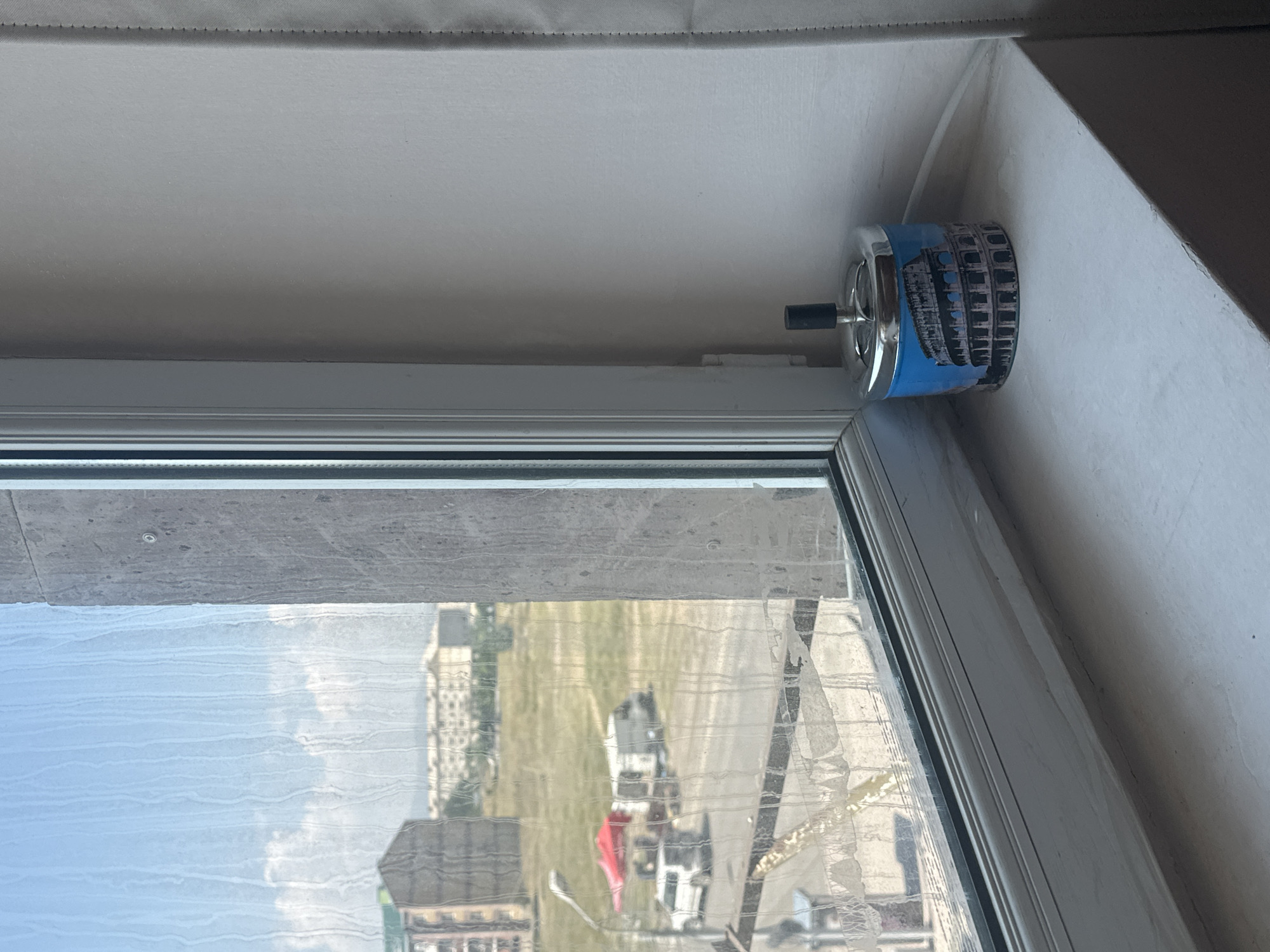 The image shows a close-up view of a windowsill in Gyumri, Armenia. On the windowsill, there is a decorative ashtray featuring an image of the Colosseum. To the left, a window looks out onto a street scene with buildings, vehicles, and a red tent. The windowpane is dirty, with streaks of water or condensation visible. The wall to the right of the windowsill is plain and white, with a section of a curtain visible on the far right edge of the frame. The overall scene suggests an indoor setting with a view of the city outside.