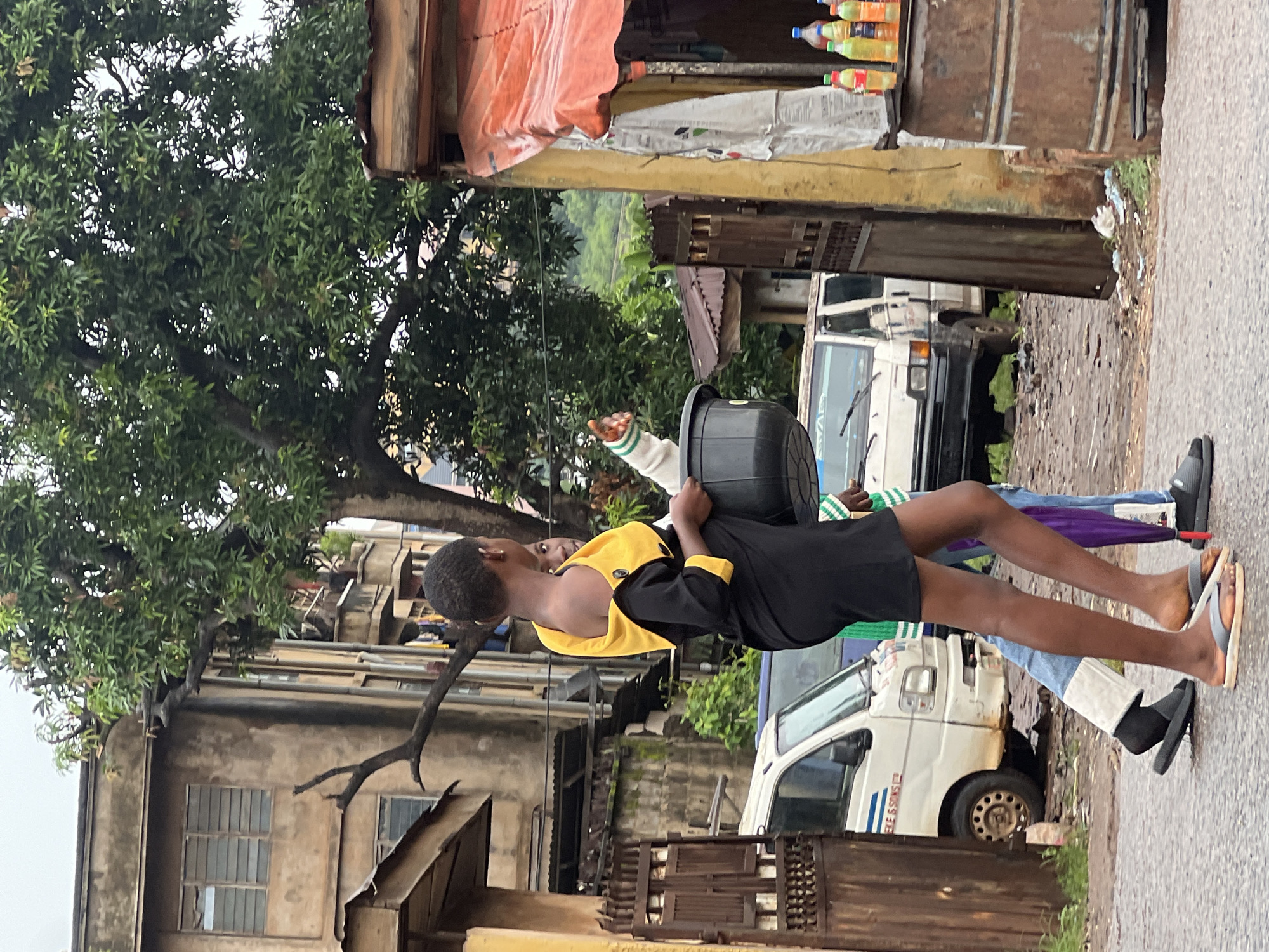Two young people are walking down a street in Nkpor, Nigeria. The person in the foreground is wearing a black dress with yellow accents and is carrying a black bucket. They are looking to the right and pointing with their left hand. The second person is partially visible and is wearing blue jeans and a green and white striped top. They are carrying a purple object.
The street is paved and appears wet, suggesting it may have rained recently. There are several vehicles on the street, including a white van with "EKE & SONS" written on the side. Buildings with weathered facades line the street, and a large tree with lush green foliage is prominent in the background. A market stall with various items, including bottles of drinks, is situated on the right side of the image. The overall atmosphere is that of a bustling urban environment.