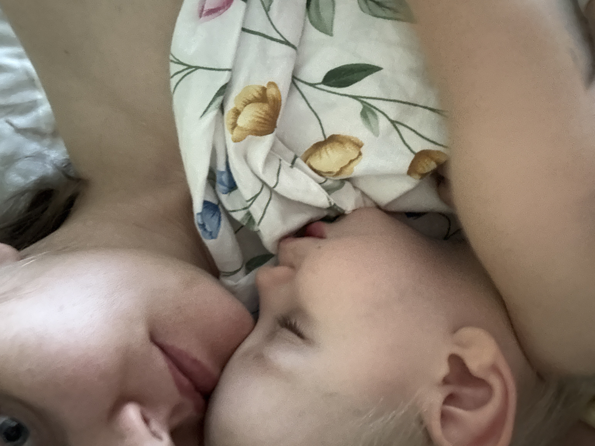 An intimate, close-up image captures an adult and a child lying cheek to cheek, embodying a moment of tenderness and closeness. The adult, positioned on the left, has a partially visible face with a gentle expression on their lips, and one eye slightly visible. On the right, the child, with light-colored hair, appears to be sleeping or resting peacefully, with their eyes closed or half-closed, and lips slightly parted.
The subjects are nestled against soft bedding. A floral-patterned fabric, likely a blanket or sheet, with delicate yellow and blue flowers and green leaves, is visible between and around them. Another lighter-toned fabric is present on the far left. The soft, diffused lighting suggests an indoor setting, likely a bed, and contributes to the serene and affectionate atmosphere.
The image powerfully conveys emotions of love, comfort, and security, characteristic of a parent-child bond. There is no visible text. This tender scene was captured in Chaikovsky, Russia.