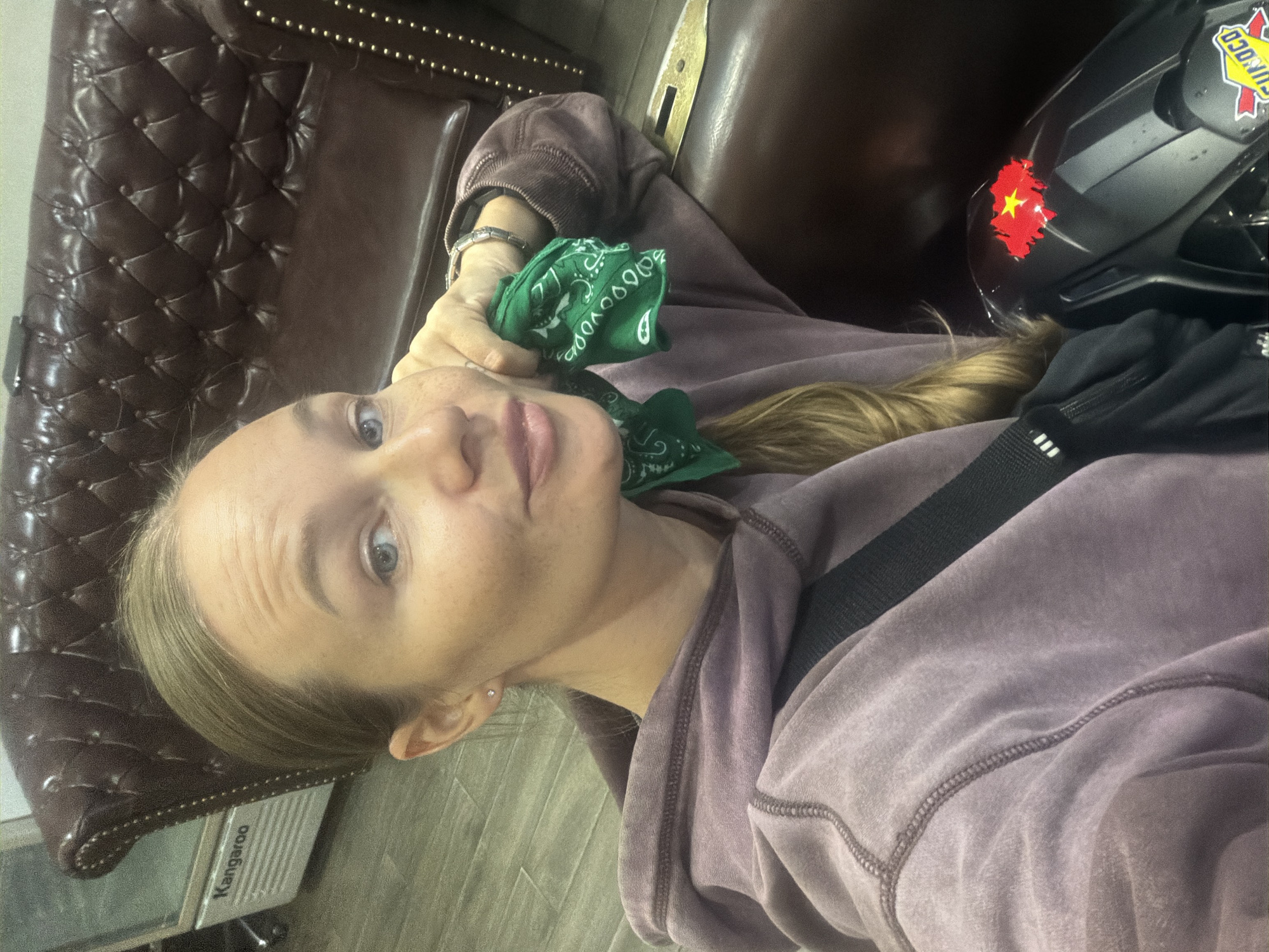The image shows a woman with blonde hair pulled back, wearing a light purple hooded sweatshirt, taking a selfie. She is holding a green bandana with a white paisley pattern to her cheek. She is sitting in what appears to be a dark leather armchair, with the back of the chair visible behind her. To the left of the armchair, a white refrigerator or cooler with the word "Kangaroo" printed on it is visible. To the right of the woman, a portion of a black motorcycle fender is visible, adorned with a red sticker depicting the shape of Vietnam with a yellow star. A yellow and red "Sunoco" logo is also visible on the motorcycle. The floor appears to be tiled. The lighting suggests an indoor setting, possibly a waiting area or a shop.
