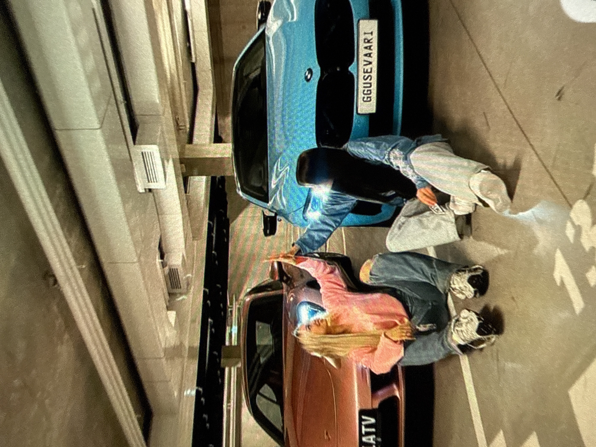Here's a detailed description of the image:
**Scene:** An indoor parking garage.
**Objects:** Two cars are parked side by side. One is light blue with a license plate that reads "GGUSEVAARI". The other is orange, with a partial license plate reading "LATV." Parking space lines are visible on the ground.
**People:** Two people are present, each leaning on one of the cars. One person is wearing a pink top and jeans, the other is wearing a blue jacket and grey pants. Their actions suggest they might be posing for a photo.
**Location:** The specific city is unknown based on the information provided in the image.