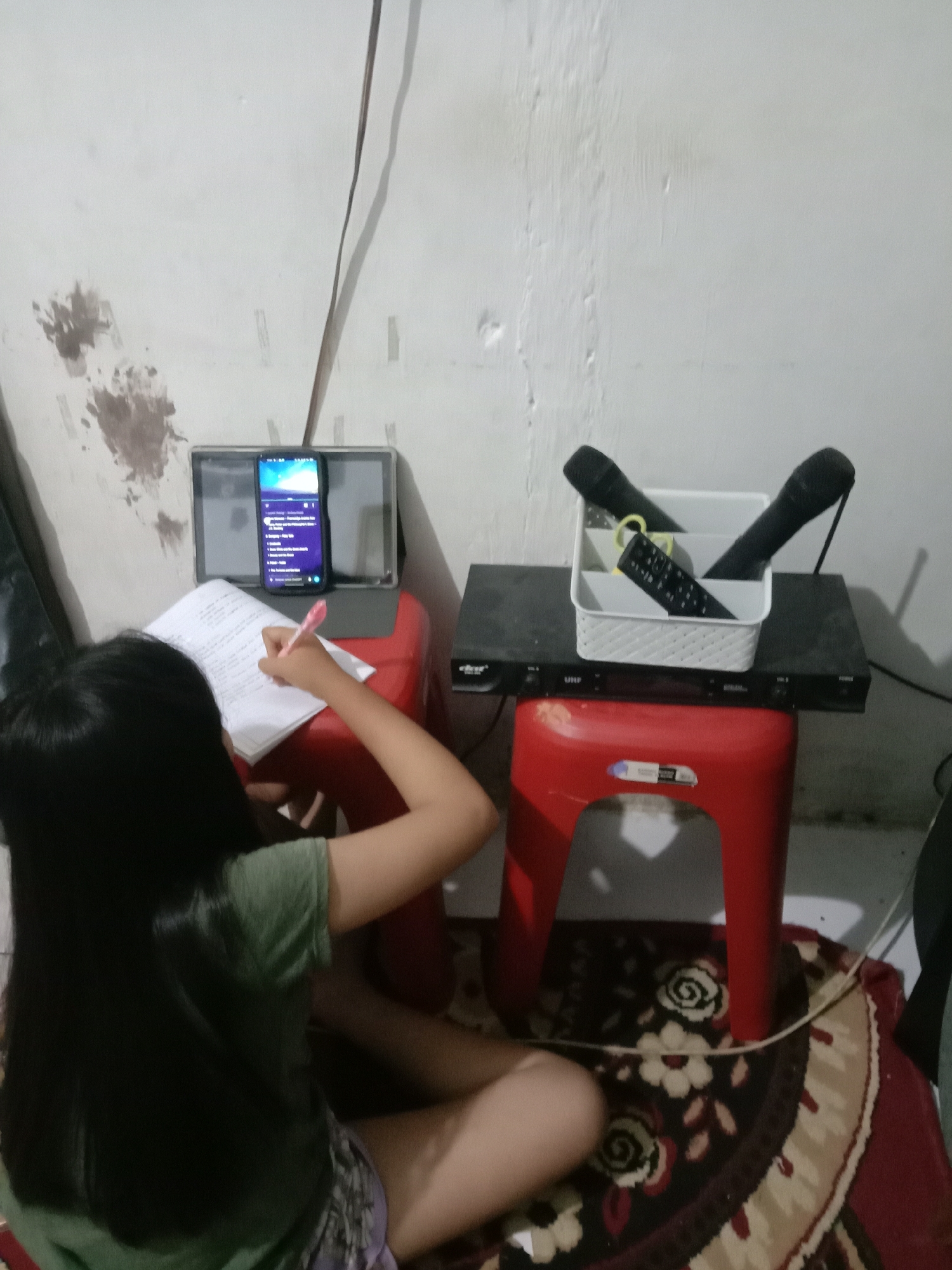 In Kwandang, Indonesia, a young girl sits on a red plastic stool, focused on writing in a notebook. She holds a pink pen in her right hand, her left hand resting on the notebook. She is wearing a green t-shirt and patterned shorts. In front of her, on another red stool, is a black DVD player with two microphones and a remote control in a white organizer on top. A tablet and smartphone are placed behind the notebook, with the smartphone displaying a playlist. The scene is indoors, with a textured white wall in the background and a patterned rug on the floor.