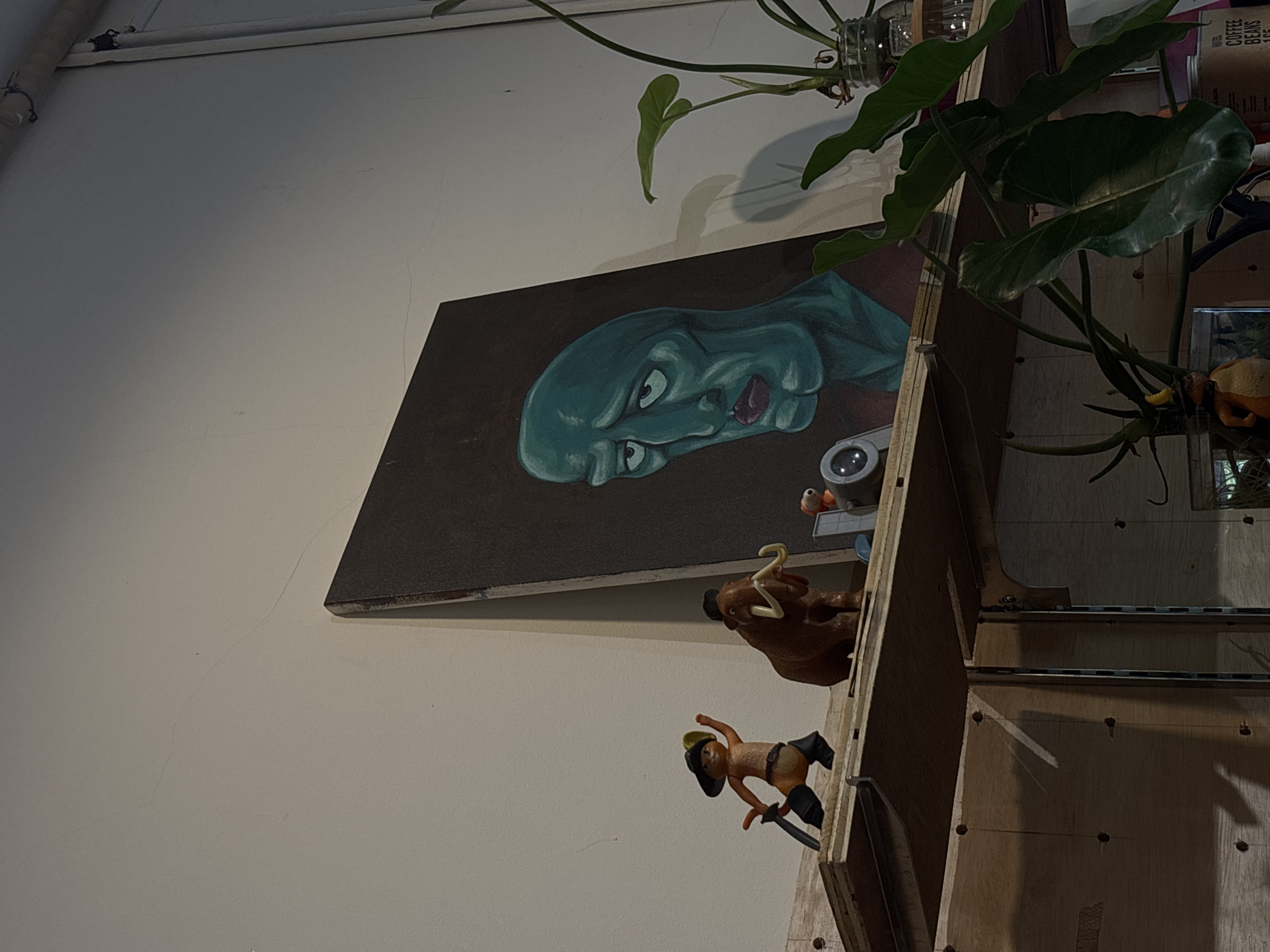 The image depicts a shelf displaying various decorative items. On the left, a small figurine of Puss in Boots is posed with a sword. Next to it stands a brown figurine of a woolly mammoth with tusks. To the right of the mammoth, a painting of the SpongeBob SquarePants character Squidward Tentacles, depicted in a dramatic and melancholic pose, is leaning against the wall. In front of the painting, a small orange figure with a yellow hat is visible, along with a circular device. A plant with large green leaves in a glass jar is situated to the right of the painting. The shelf itself is made of wood and is supported by metal brackets. The wall behind the shelf appears to be painted white and shows some minor imperfections. The overall impression is that of a curated display of eclectic items, possibly in a home or a small shop. The image does not contain enough information to determine its location in Semarang, Indonesia.