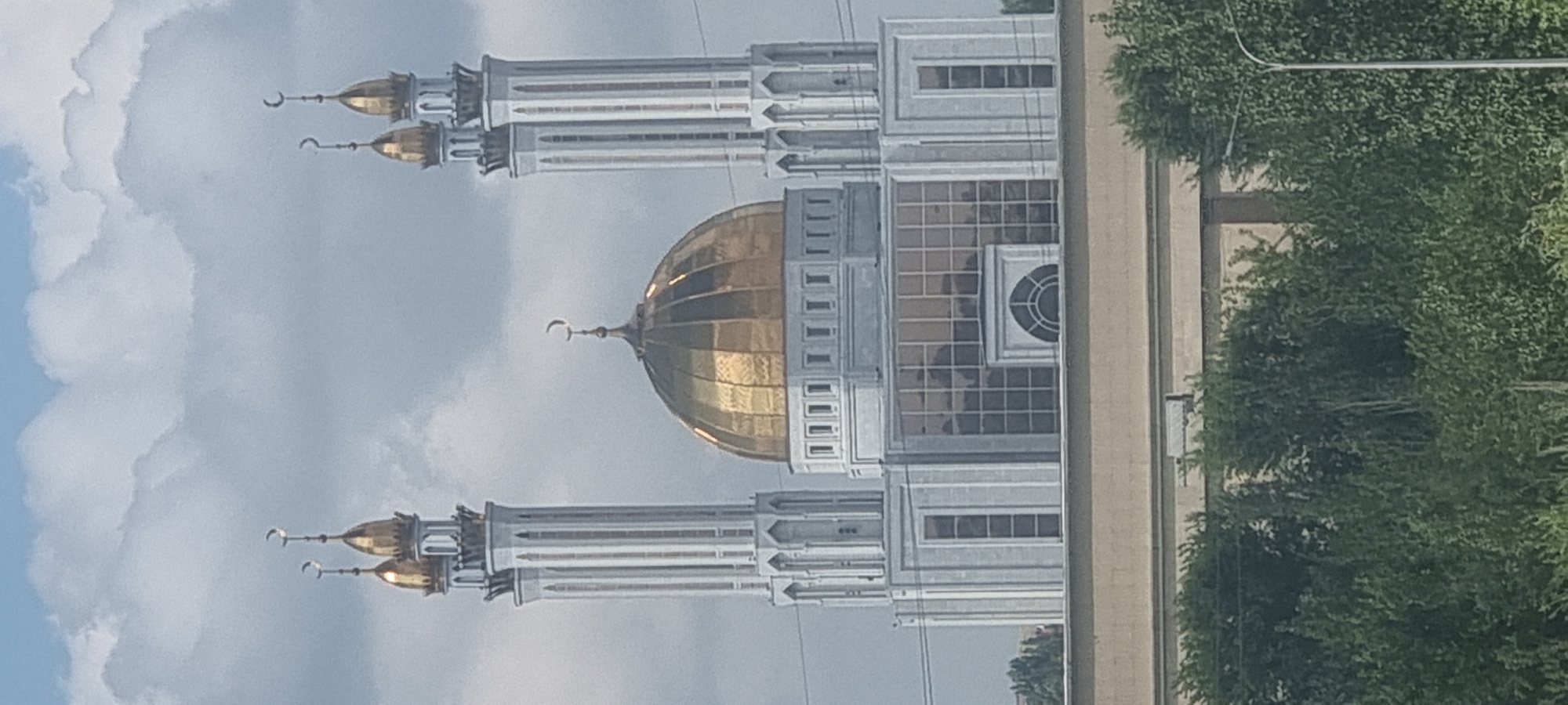 The image shows a mosque in the city of Aktobe, Kazakhstan. The mosque features a large golden dome and two tall white minarets, each topped with a golden crescent moon. The building's exterior is predominantly white with decorative elements and large glass windows. In the foreground, there is a dense green foliage of trees, and a beige-colored structure is visible beneath the mosque. The sky above is overcast with clouds. No people or specific actions are depicted in the scene.