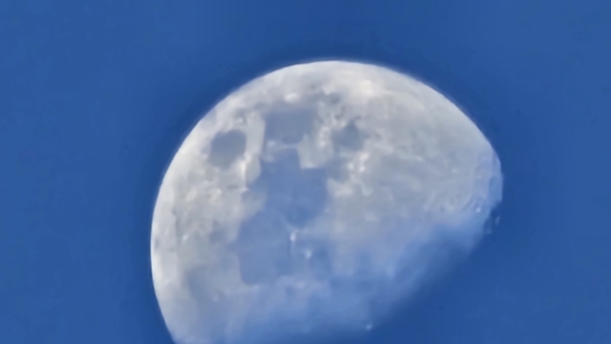 A half-illuminated moon is captured against a clear, bright blue sky. The moon's surface exhibits visible craters and maria, creating a textured appearance. The lighting suggests it is daytime, possibly late afternoon or early morning, when the moon can be seen in the sky. The image is a close-up shot, focusing solely on the celestial body and its backdrop. There is no indication of people, specific activities, or explicit location cues beyond the general setting of the sky. No text is visible in the image.