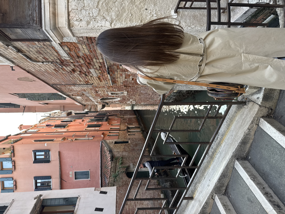 The image depicts a scene along a narrow canal in Venice, Italy, viewed from a stone bridge. In the immediate foreground, a person with long, straight brown hair, seen from behind, wears a light beige long trench coat and a circular brown patterned shoulder bag with a tan strap. Their hands are raised, positioned as if holding a device to capture a photograph of the canal and the buildings opposite. To the left, a flight of concrete steps leads down, bordered by a dark metal railing with geometric rectangular patterns. Below the bridge, on the canal, a gondola is propelled by a male gondolier. He stands in the black gondola, facing away from the camera, wearing a dark vest over a striped shirt and dark trousers, and operates a long oar. The canal water reflects sunlight. Along the canal banks, various Venetian buildings rise. On the left, a light-colored building is partially visible, adjacent to a prominent multi-story structure with reddish-pink walls and numerous windows, some fitted with dark shutters. At the base of this building is a smaller, older brick structure with a tiled roof. On the right, the bridge's stone foundation gives way to a brick building featuring exposed brickwork, light plaster sections, and arched architectural details, continuing down the canal.