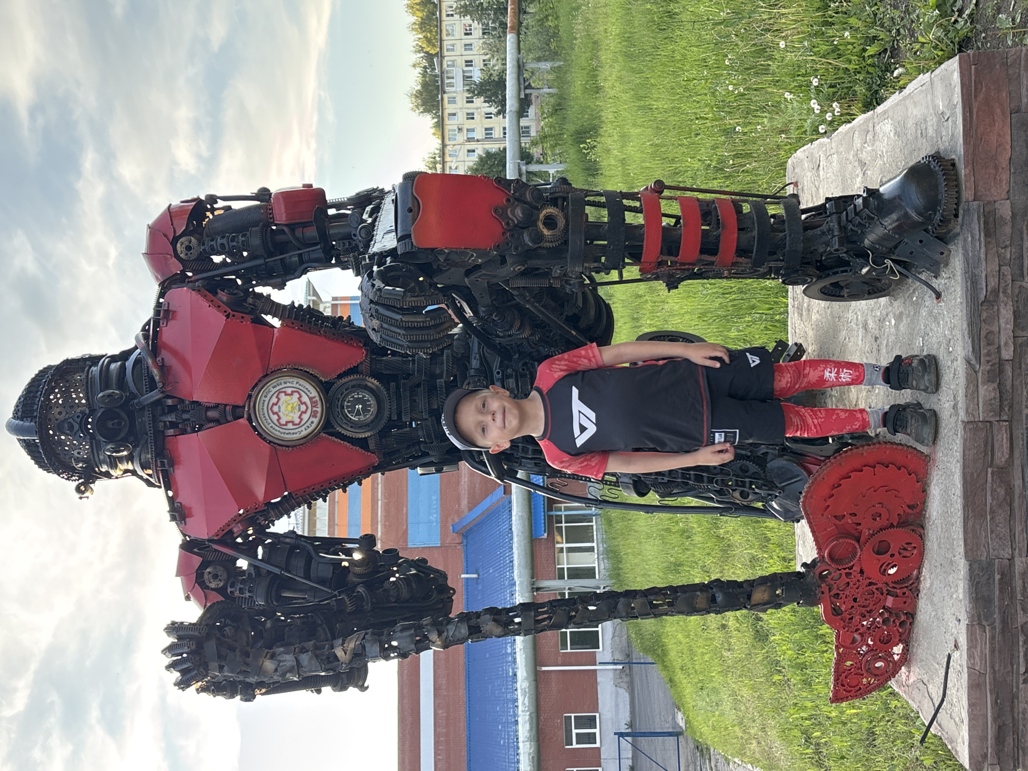 A young boy poses proudly next to a large, intricate robot sculpture made from recycled metal parts. The scene is set outdoors, likely in a public area of Votkinsk, Russia, given the contextual information.
The boy, fair-skinned with light hair, wears a baseball cap, a black t-shirt with a white stylized 'V' or triangular emblem, black shorts, red socks featuring white characters, and black sneakers. He stands on the right side of the imposing robot, looking towards the camera with a slight smile.
The robot sculpture is a remarkable piece of art, predominantly red and black. It is constructed from a multitude of discarded mechanical components, including gears, chains, engine parts, and metal sheeting, giving it a robust, industrial, and almost fierce aesthetic. Its left arm (from the viewer's perspective) ends in a large, red, axe-like structure also crafted from gears and metal. On the robot's chest, a circular emblem is visible, bearing text in Cyrillic which reads, "ФГБУ N 80 МЧС России" (Federal State Budgetary Institution No. 80 EMERCOM of Russia), with "спасенные" (rescued) and "VOTKINSK" also discernible.
In the background, there's green grass with some white dandelions, and in the distance, various buildings are visible, including what appears to be an apartment block and structures with blue roofs. The sky above is partly cloudy, suggesting it's daytime, possibly late afternoon or early evening with soft lighting. The robot stands on a concrete plinth, which has a reddish-brown brick facade. The white characters on the boy's red socks appear to be the Chinese characters "休闲" (xiūxián), meaning "casual" or "leisure."
