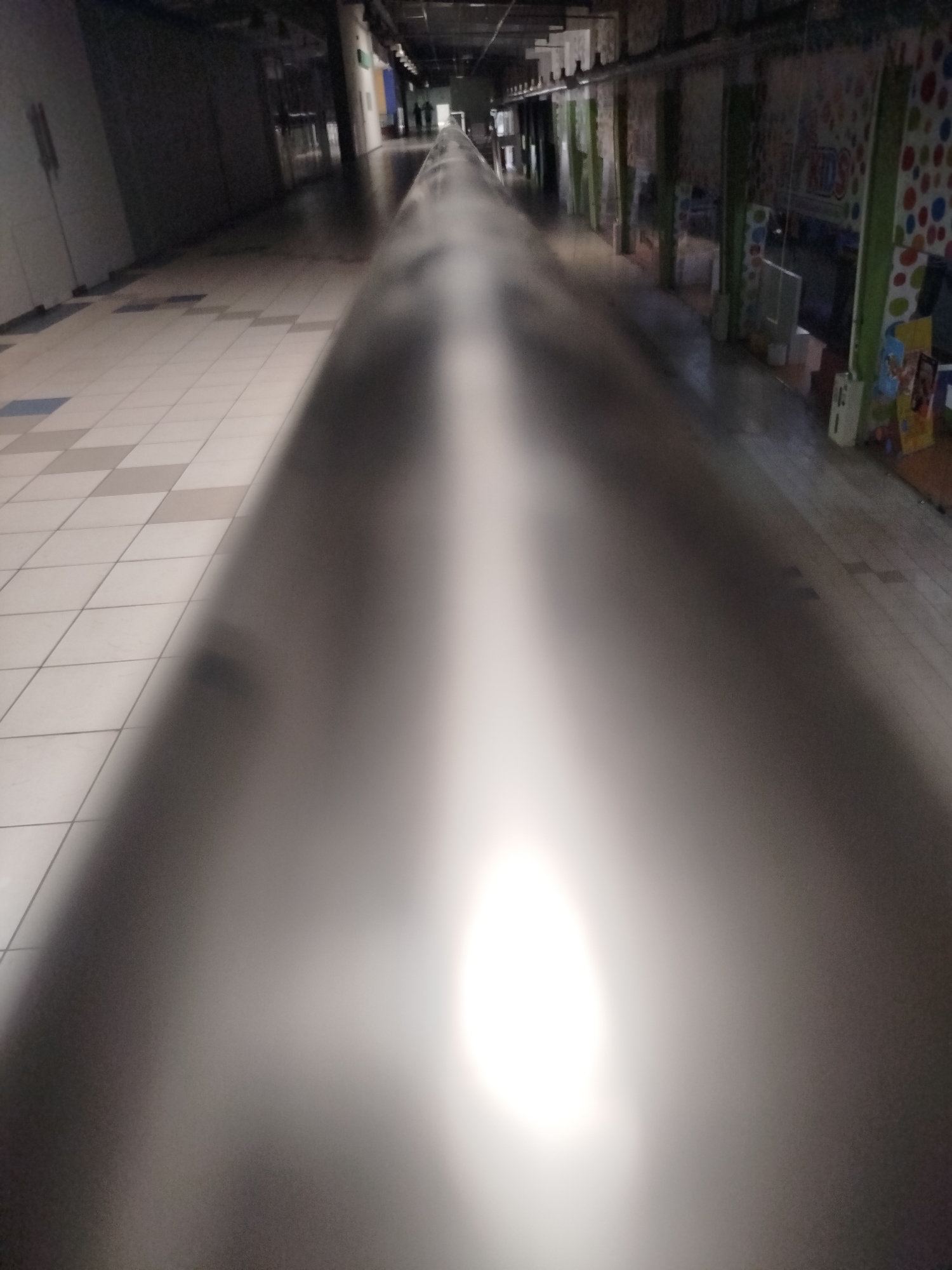 The image shows a long, dimly lit hallway or corridor, likely within a commercial or public building. The perspective is from a low angle, looking down the length of the corridor. On the left side, a tiled floor is visible, with a pattern of light and dark square tiles. The right side of the corridor is occupied by what appears to be a railing or a long, dark, reflective surface.
Further down the corridor, the walls are lined with what seem to be storefronts or rooms, some with green frames around their entrances. One storefront on the right has a colorful decoration with red, blue, and yellow dots, and the word "KIDS" is visible.
The overall atmosphere is somewhat desolate and empty. There are no clear signs of activity or people in the immediate foreground. The lighting is dim and uneven, with a bright, blown-out area in the center of the image, suggesting a light source or reflection. The image quality is poor, with significant blur and noise, making it difficult to discern fine details. The location is not specified in the image itself, and therefore, its presence in Kremenchuk, Ukraine, cannot be determined from the visual information provided.