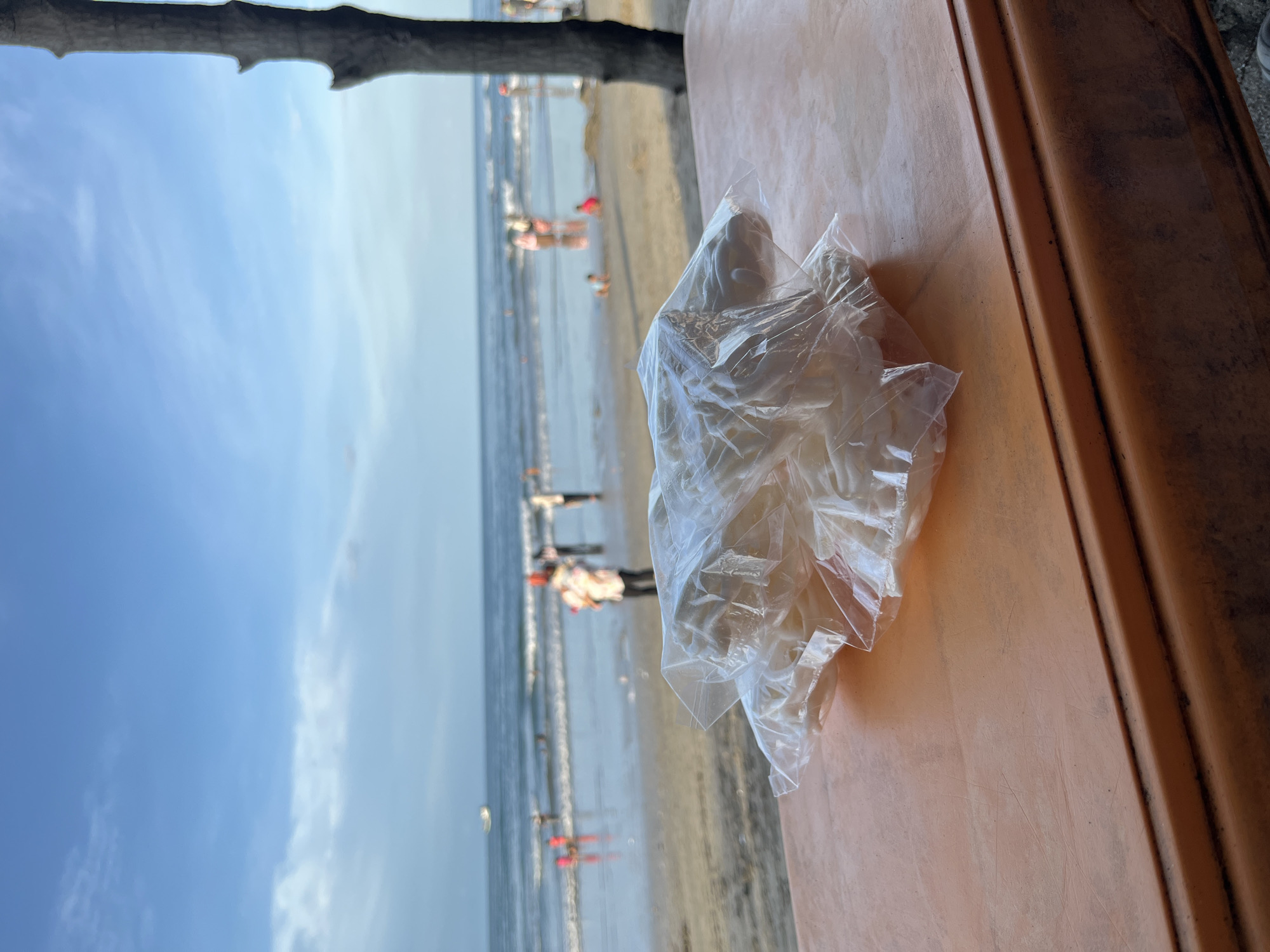 Viewed from a slightly elevated position in Lhokseumawe, Indonesia, a partially obscured, slightly blurry scene presents an outdoor setting where the sky is partly visible. An unknown object in a clear plastic bag sits prominently on a peach-colored surface in the foreground. The beach, water, and horizon line are visible in the background. Several indistinct figures can be observed in the water and on the beach. The horizon line is slightly above the image's vertical midpoint.