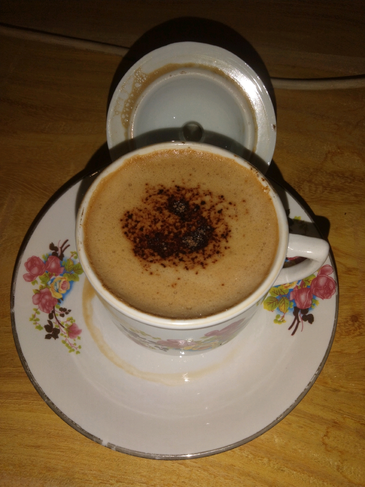 A close-up, top-down shot of a cup of coffee with frothy, light brown crema and a sprinkle of dark coffee grounds on top, rests on a saucer with a floral pattern of pink, red, and blue flowers. The cup and saucer are white with a metallic silver rim. A white lid, also with a metallic silver rim, is placed behind the cup, slightly ajar, revealing a similar metallic silver interior rim. The coffee and ceramic set are placed on a light brown, wooden surface, which appears to be a table. The lighting is dim, creating a shadow that obscures the background. The image was taken in Merakurak, Indonesia.