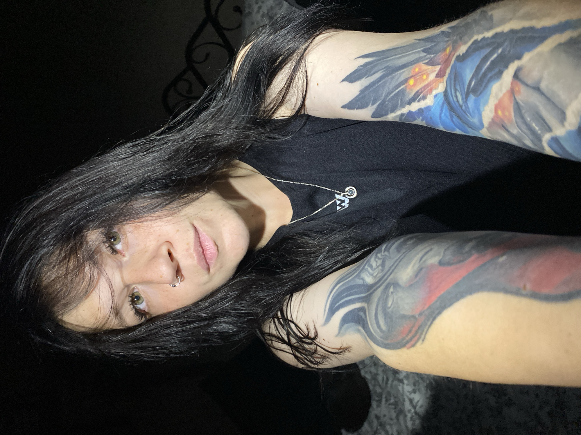 A young woman with long, dark hair and green eyes is pictured in a close-up, overhead shot. She is wearing a black tank top with a silver necklace featuring a pendant with a "W" symbol and a green stone. She has a nose ring and several tattoos on her arms. One tattoo on her left arm appears to be a red and black abstract design, while the tattoo on her right arm depicts a blue and grey feathered creature with orange accents. The background is dark and out of focus, with a hint of what appears to be a headboard of a bed. The lighting is dim, creating shadows on her face and arms. This image was not taken in Chaikovsky, Russia.