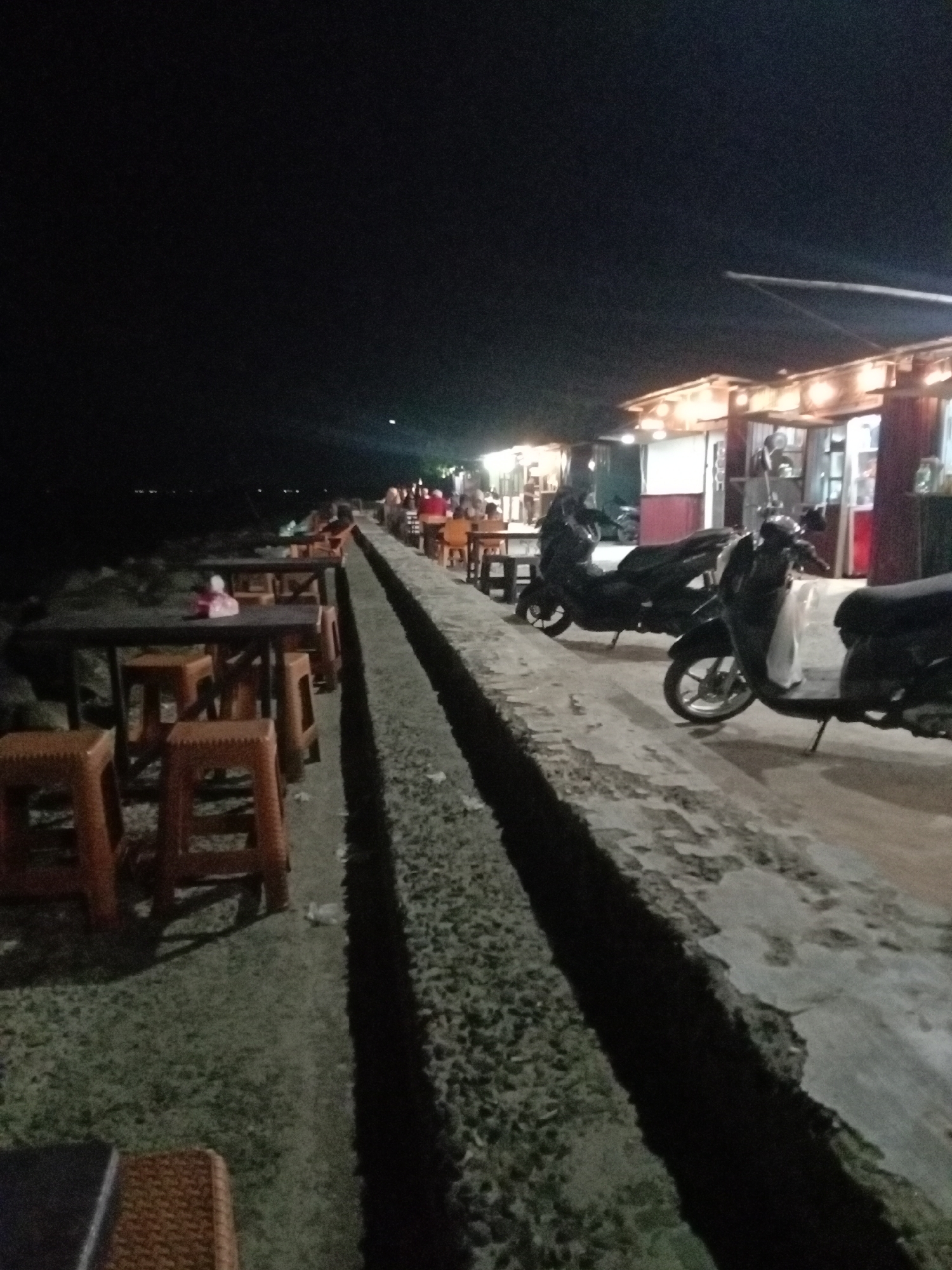 The image depicts a nighttime scene at a bustling outdoor dining area, likely a series of food stalls or cafes, along what appears to be a coastal path or promenade in Lhokseumawe, Indonesia.
The foreground is dominated by a long, concrete pathway that is bisected lengthwise by a narrow, rough-hewn channel or drain. To the left of this channel, several rows of dark tables paired with orange plastic stools are set up. A red tissue box is visible on one of the tables in the foreground. Beyond these tables, the area quickly fades into darkness, hinting at a large body of water or an unlit expanse. Distant figures of people can be seen seated at some of these tables, suggesting patrons enjoying a meal or conversation.
On the right side of the concrete channel, a line of small, brightly lit food stalls or shops extends into the distance. These structures feature simple roofs and emit a warm, inviting glow, illuminating their surroundings. Several scooters and motorcycles are parked in front of these stalls, indicating the presence of customers. People are also visible near and inside these stalls, engaged in various activities like serving or dining.
The overall scene conveys a casual and social atmosphere typical of an evening gathering spot. The time of day is clearly night, with the sky appearing dark and the artificial lighting from the stalls being the primary source of illumination. The image has a slightly grainy quality due to the low-light conditions. No discernible text is visible.