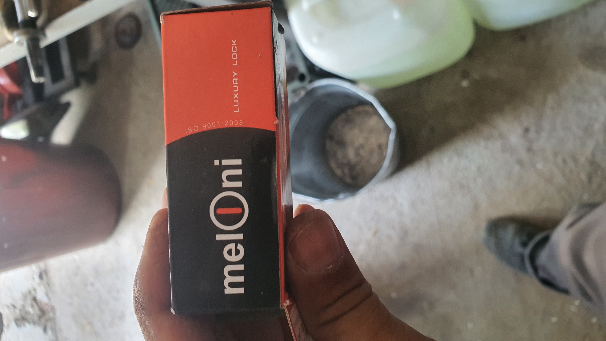 A close-up, eye-level shot shows a person's hand holding a black and orange box. The box has white text that reads "melOni" with a circular design in the center of the "O", and on the right side, text reads "ISO 9001:2008 LUXURY LOCK". The background is blurred but appears to be an indoor setting with a concrete floor, a metallic bucket, and large, translucent containers. A person's feet wearing dark shoes and pants are visible at the bottom of the frame, suggesting they are standing. The lighting is natural and casts a shadow.