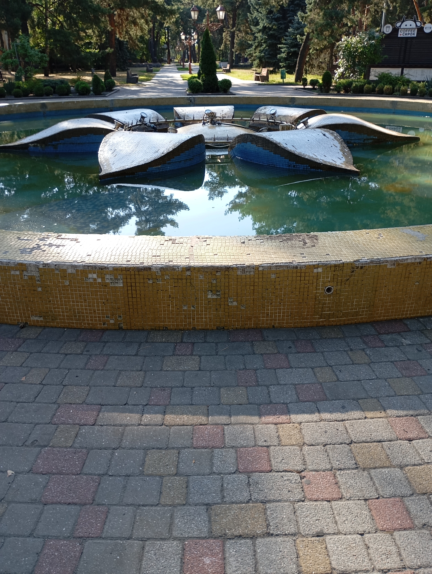 In Horishni Plavni, Ukraine, a partially submerged fountain features petal-shaped structures covered in white and blue tiles, resting in teal-colored water. The fountain's circular rim is adorned with aged yellow mosaic tiles, some of which are chipped and missing. In the foreground, a pathway constructed from interlocking grey, pink, and yellow paving stones is visible, with a clear shadow cast diagonally across it. Beyond the fountain, a park setting unfolds, complete with mature trees, manicured bushes, park benches, and lampposts lining a paved walkway. In the background, a sign for "COFFEE & SIROK" is mounted on a dark structure. The overall scene depicts a public park area with a prominent, albeit weathered, water feature.