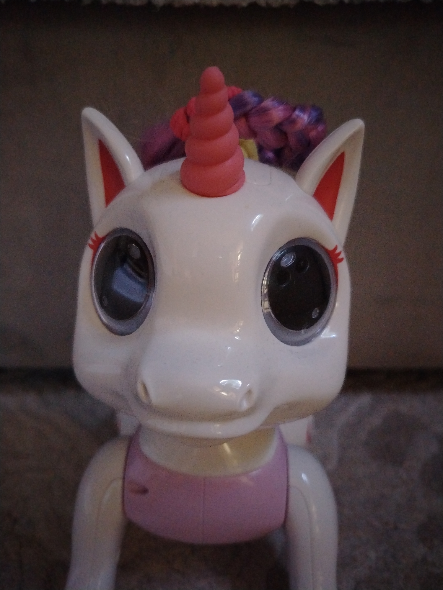 The image shows a close-up view of a white plastic toy unicorn. The unicorn has large, round black eyes, a pink horn, and a pink and purple braided mane. The unicorn is wearing a pink top. The background is blurry and appears to be a textured surface, potentially a wall. The provided location is Kaduy, Russia, but there's no indication of Kaduy or any specific location within it in this image. The toy is the primary subject, filling much of the frame. There are no people visible in the image.