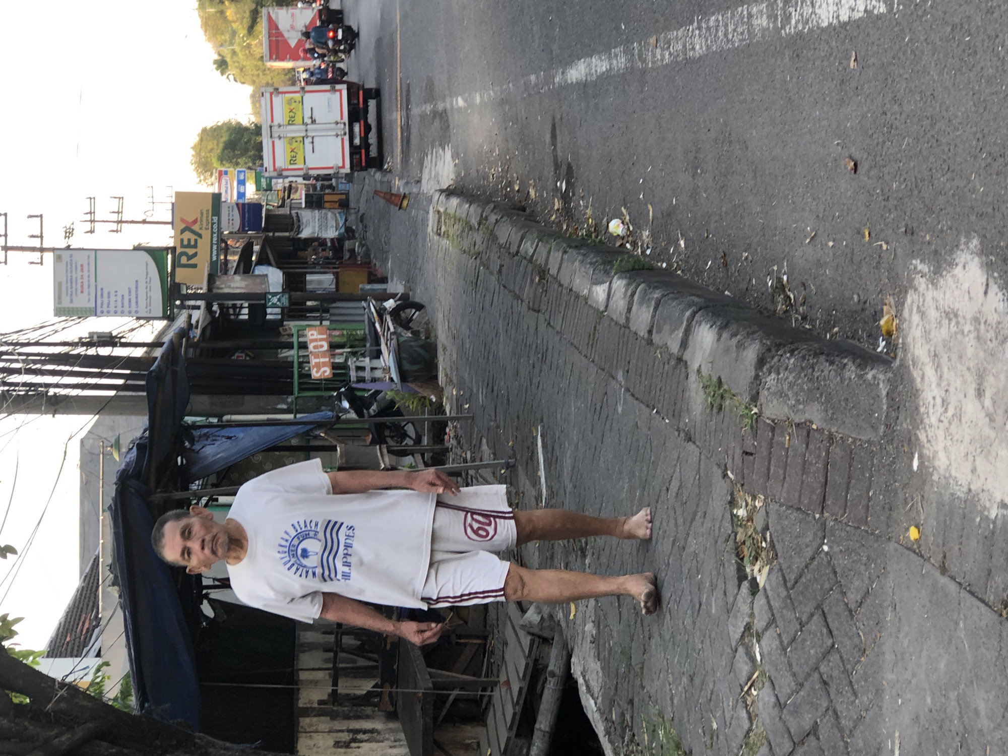An elderly East Asian man, wearing a white t-shirt with blue text and graphics that read "MATABUGKAY BEACH PHILIPPINES" and white shorts with red stripes and the number "60" in a red circle, walks barefoot along a paved sidewalk on a street in Ngabang, Indonesia. He is facing forward and his gaze is directed slightly downward. To his left, there are several market stalls with blue tarps and wooden tables. To his right, a paved road stretches into the distance, with a white line marking the shoulder. Several vehicles, including white box trucks and motorcycles, are visible on the road. The overall environment appears to be a commercial or street market area, with utility poles and wires visible overhead. The lighting suggests it is daytime, possibly late afternoon or early morning due to the soft, diffused light.