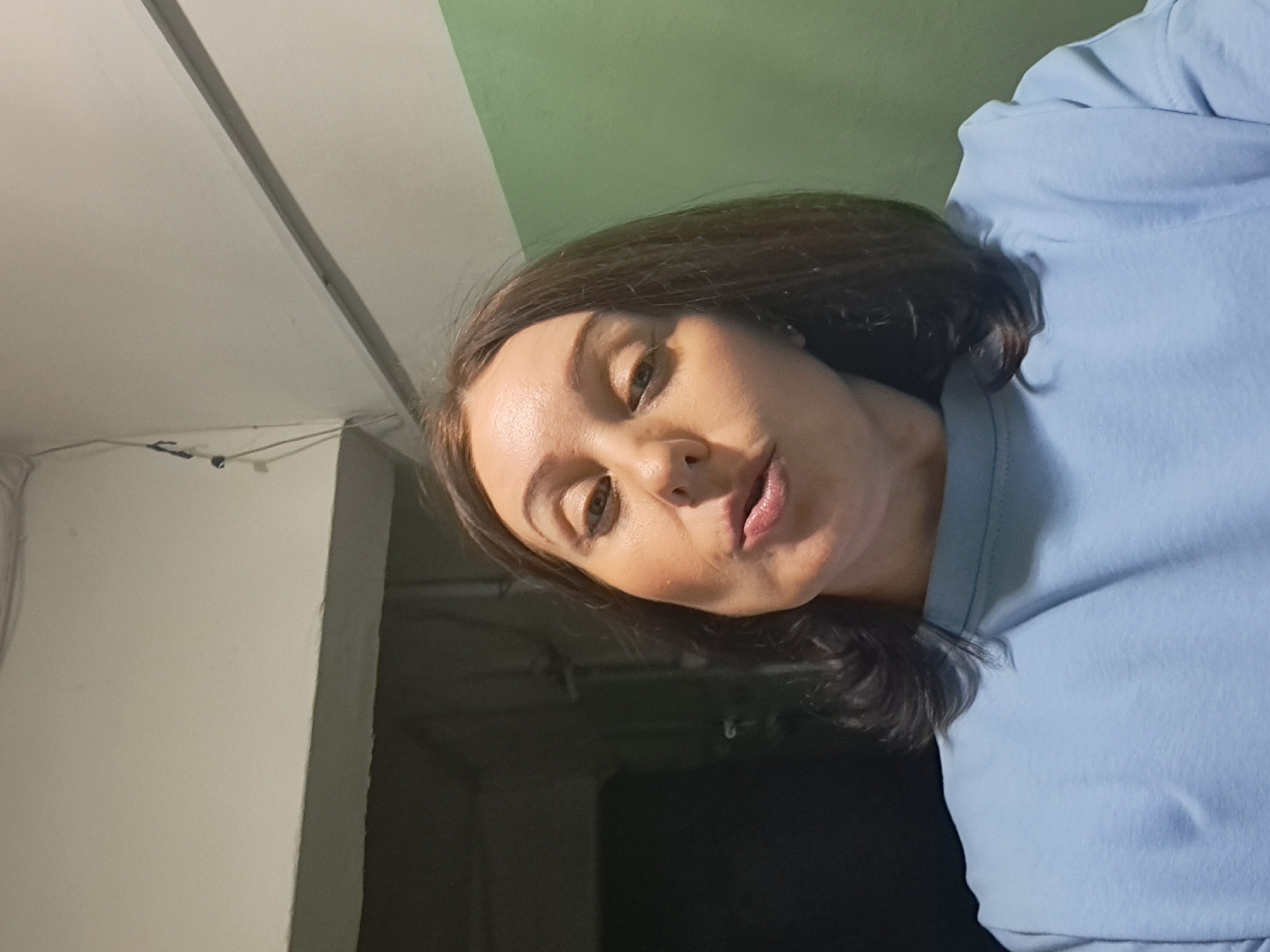 The image shows a close-up, waist-up, indoor selfie of a woman with medium-length brown hair. She is wearing a light blue sweatshirt. Her lips are slightly pursed, and her head is tilted slightly. The background appears to be a hallway or corridor with walls painted in light gray and green. There are some electrical wires visible near the ceiling. The lighting is dim. The image does not provide any specific details that would indicate the location in the city of Irkutsk, Russia.