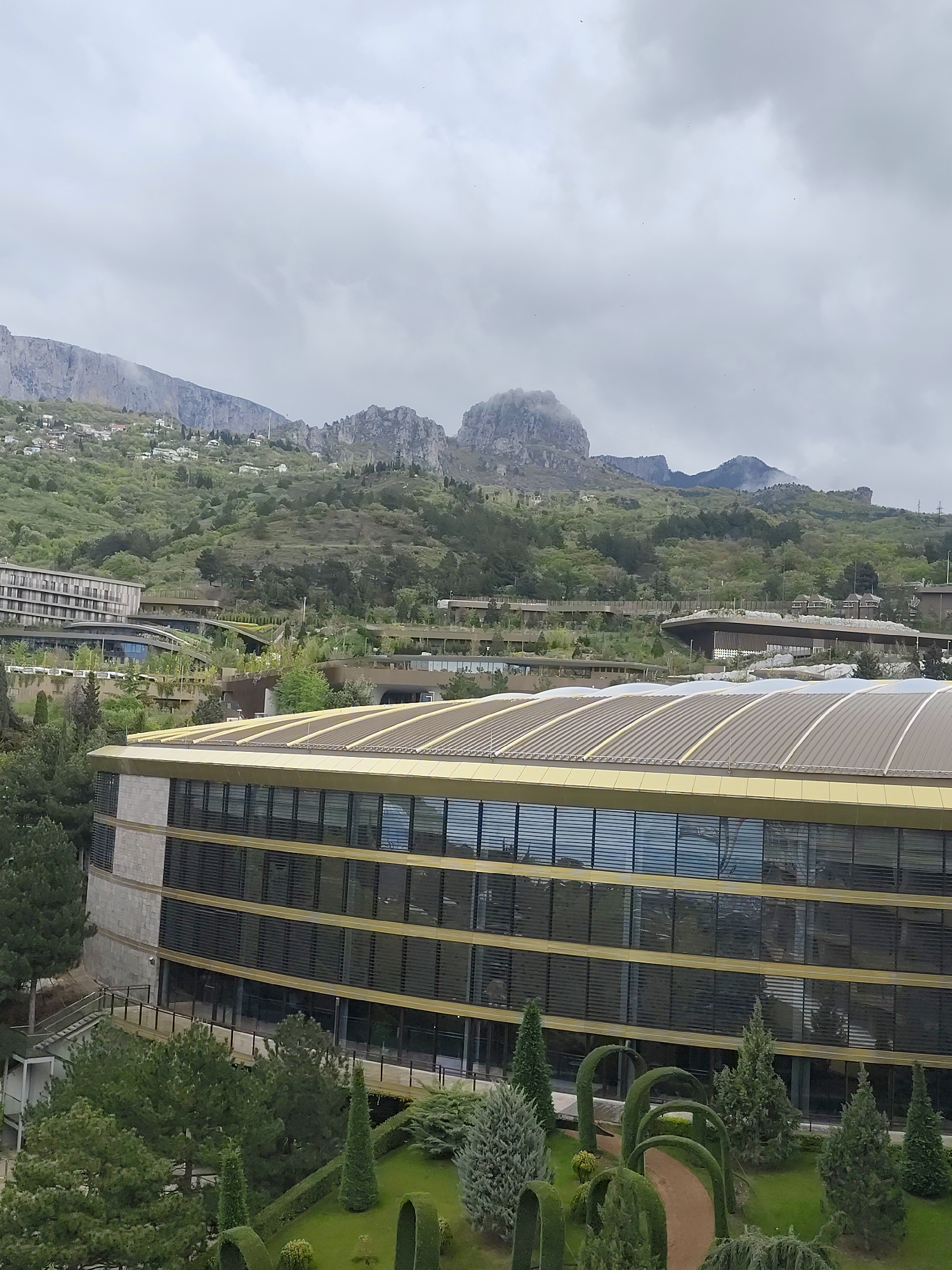 The image shows a view of a modern building with a curving facade, situated in a landscape that includes mountains in the background, likely in or near Симеизский поселковый совет, Ukraine. The building features a rounded roof, multiple rows of windows, and a golden or tan accent color. The foreground is dominated by greenery, including trees and manicured shrubs and hedges, some of which are shaped into arches, forming a garden path. In the mid-ground, there are other buildings partially visible, and the background features lush, green hills leading up to rocky mountains partly obscured by clouds. The sky is overcast.