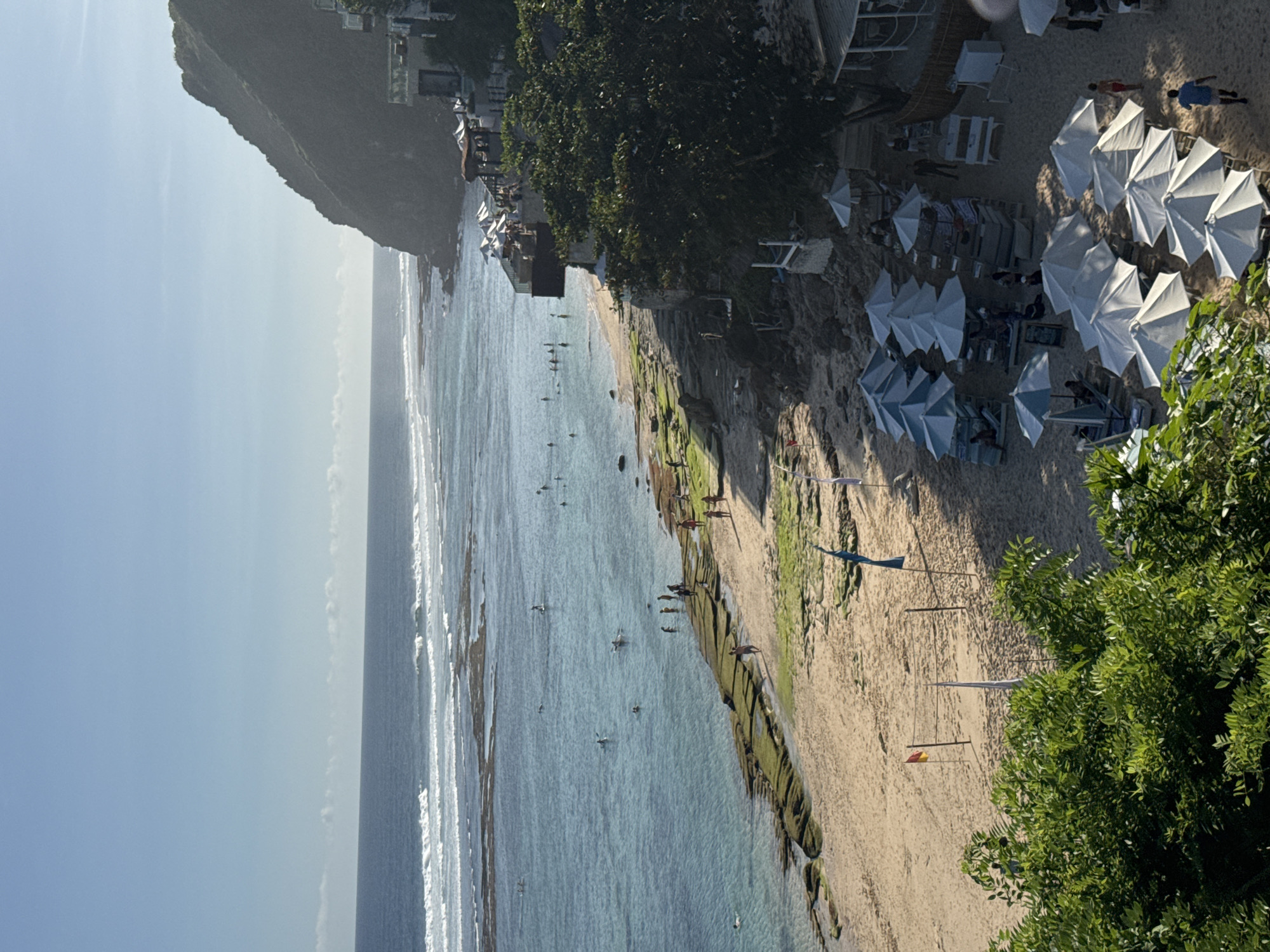 The image depicts a coastal scene in Kuta Selatan, Indonesia. The ocean extends into the distance, with visible waves and several people engaging in water activities like swimming and surfing. A beach runs along the edge of the water, marked by visible green algae-covered rock formations. Further inland, the beach transitions into a sandy area dotted with beach umbrellas and lounge chairs, indicating a resort or recreational zone. People are seen relaxing under the umbrellas. Buildings and trees line the edge of the beach, suggesting a developed coastal area backed by natural vegetation. A rocky landform or hill is also visible in the distance.