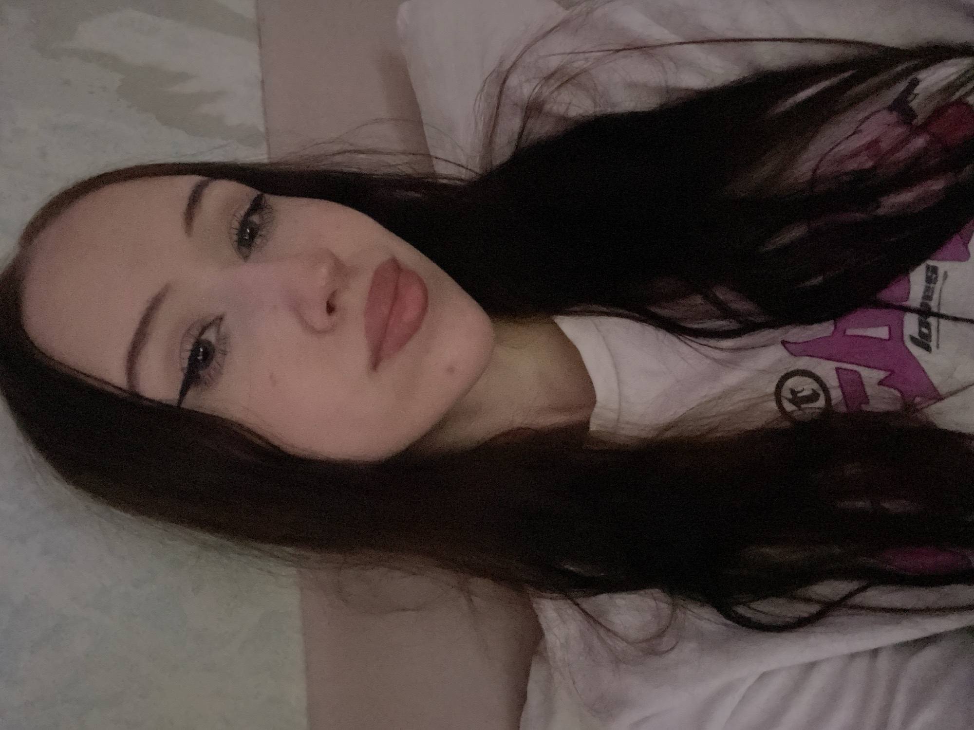 The media file is a close-up, low-angle selfie of a young person lying down, with their face tilted towards the camera. The individual has long, dark brown hair that cascades around their face and shoulders. They are wearing a white t-shirt with a graphic design that includes purple lettering and a circular logo. The background appears to be a bed with a patterned duvet cover, suggesting an indoor setting, likely a bedroom.
The person's face is the primary focus, with their eyes open and looking directly at the camera. They have noticeable eye makeup, including winged eyeliner and mascara, and their lips are painted with a light pink lipstick. The lighting is soft, and the overall mood seems relaxed, possibly indicating a casual moment. There are no explicit cues about the time of day or weather, but the indoor setting and soft lighting suggest it could be evening or a dimly lit room.
The image was captured in Votkinsk, Russia. No specific activity or event is clearly depicted beyond the act of taking a selfie. Notable details include the individual's makeup and the t-shirt's design, though the text on the t-shirt is partially obscured and difficult to read fully. The overall impression is an intimate, personal snapshot.