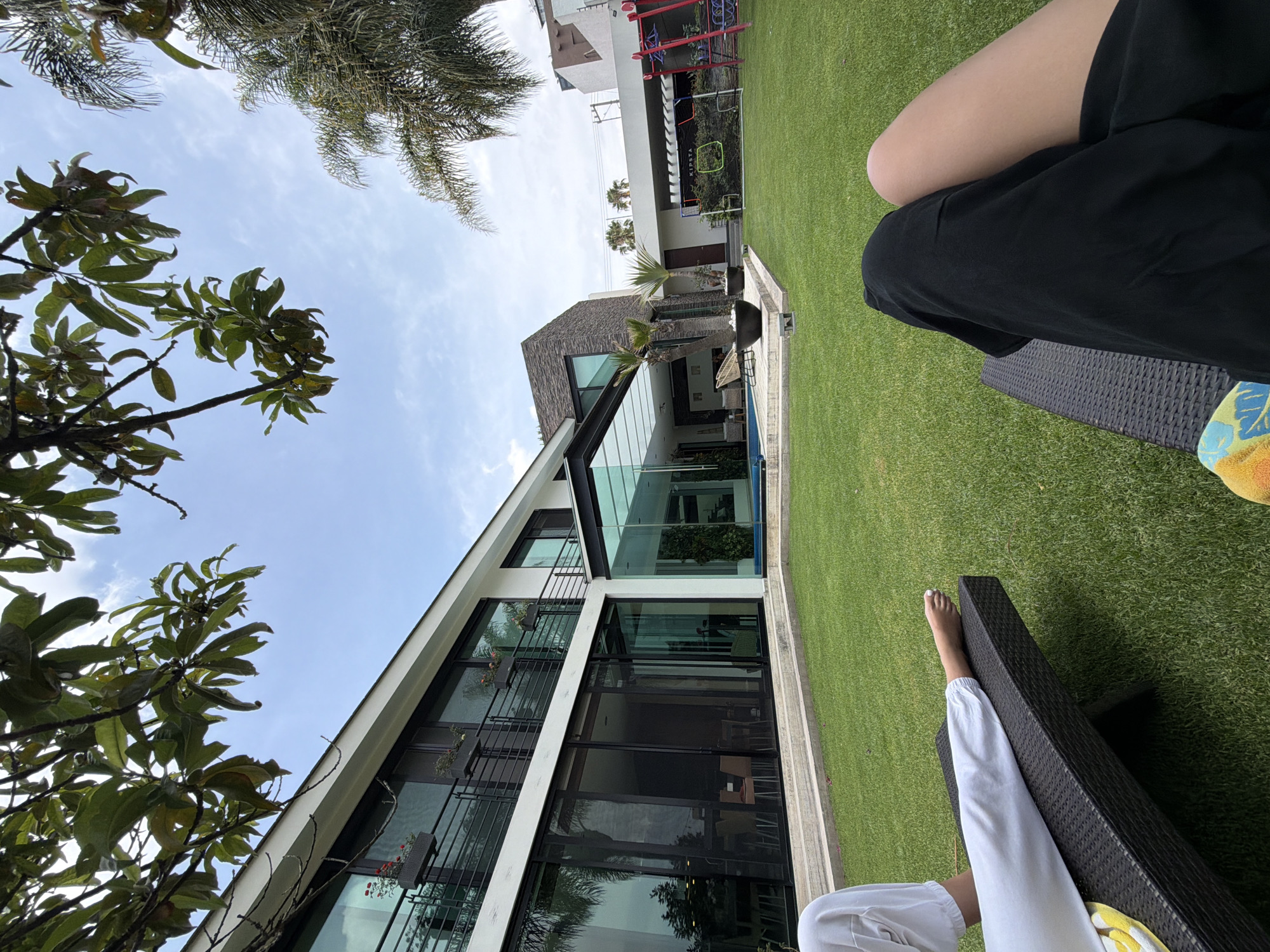 The photograph shows a person sitting on a black wicker lounge chair on a green lawn next to a modern house with large glass windows in Unknown, Unknown. Another person sits in a similar chair nearby. The modern house has several balconies with flower pots. Trees are on both sides of the building. There is a soccer goal in the distance near the house. The sky is blue with some clouds.