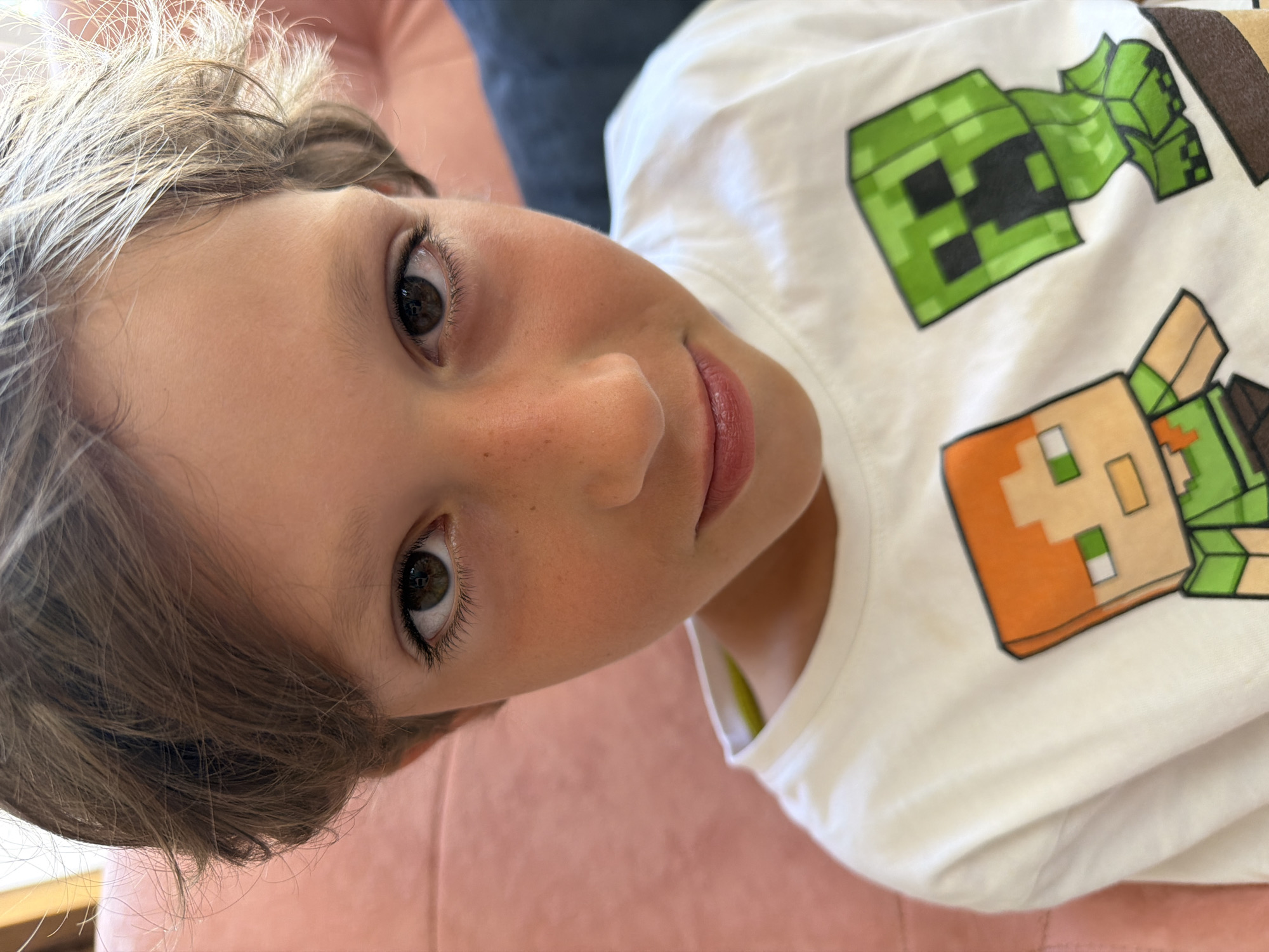 This is a close-up, high-angle shot of a young child wearing a white t-shirt featuring characters from the video game Minecraft. The child's face is turned towards the camera, with their left eye prominently visible. They have light brown hair with lighter highlights, and light skin with a scattering of freckles across their nose and cheeks. Their lips are slightly parted, revealing a hint of teeth, and they have a gentle smile. The t-shirt has a pixelated design of Minecraft characters, including a green Creeper and a character with orange and brown clothing. The background is blurred, showing a soft pink surface, possibly a sofa or chair. The lighting appears to be natural and bright, suggesting an indoor setting during the daytime. The child's expression conveys a sense of warmth and innocence. No visible text is present other than the design on the t-shirt.